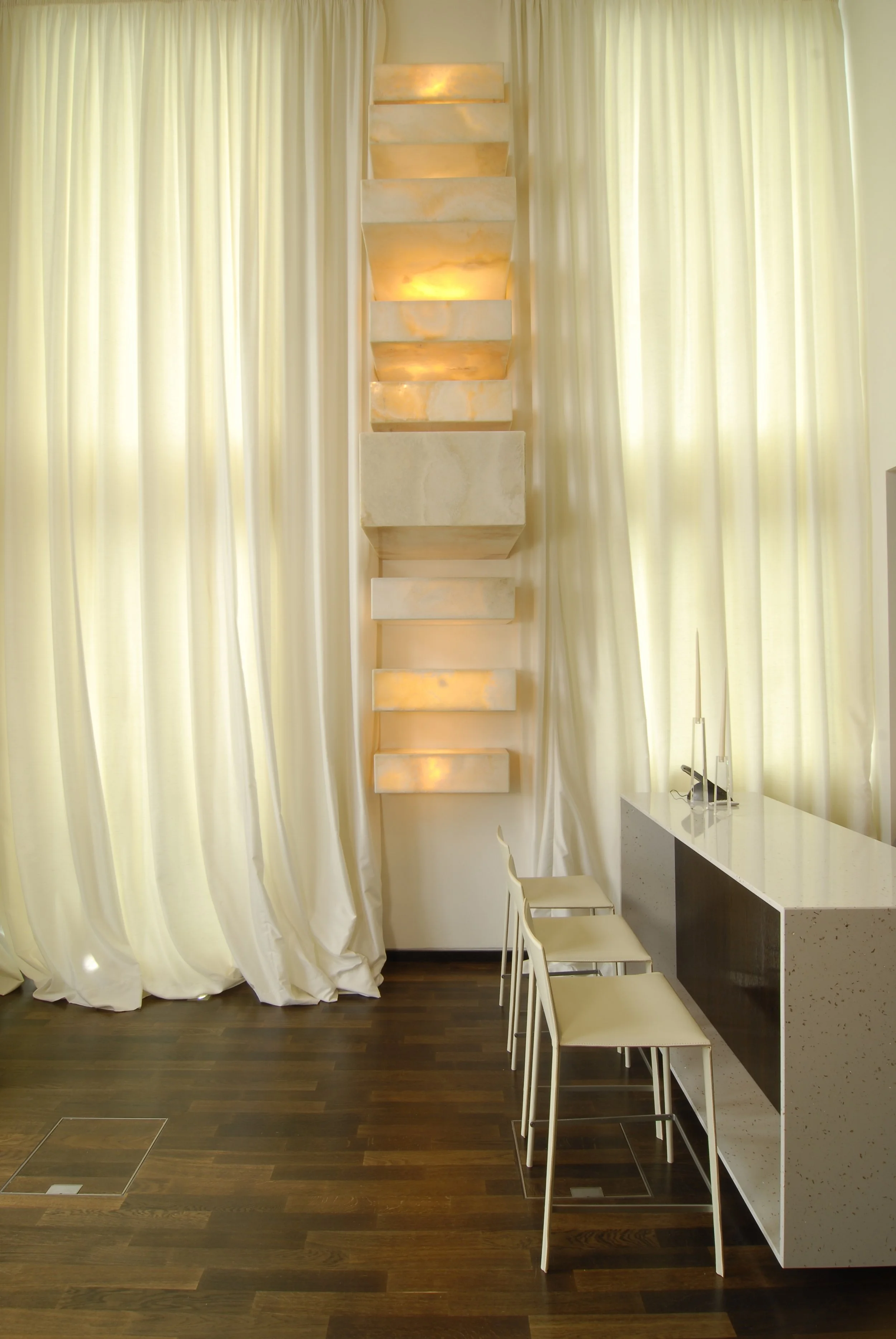 Elegantes Interieur mit weißen Vorhängen, einer aufsteigenden, beleuchteten Wanddekoration aus Marmorblöcken und einem weißen Steharbeitsplatz mit Barhockern.