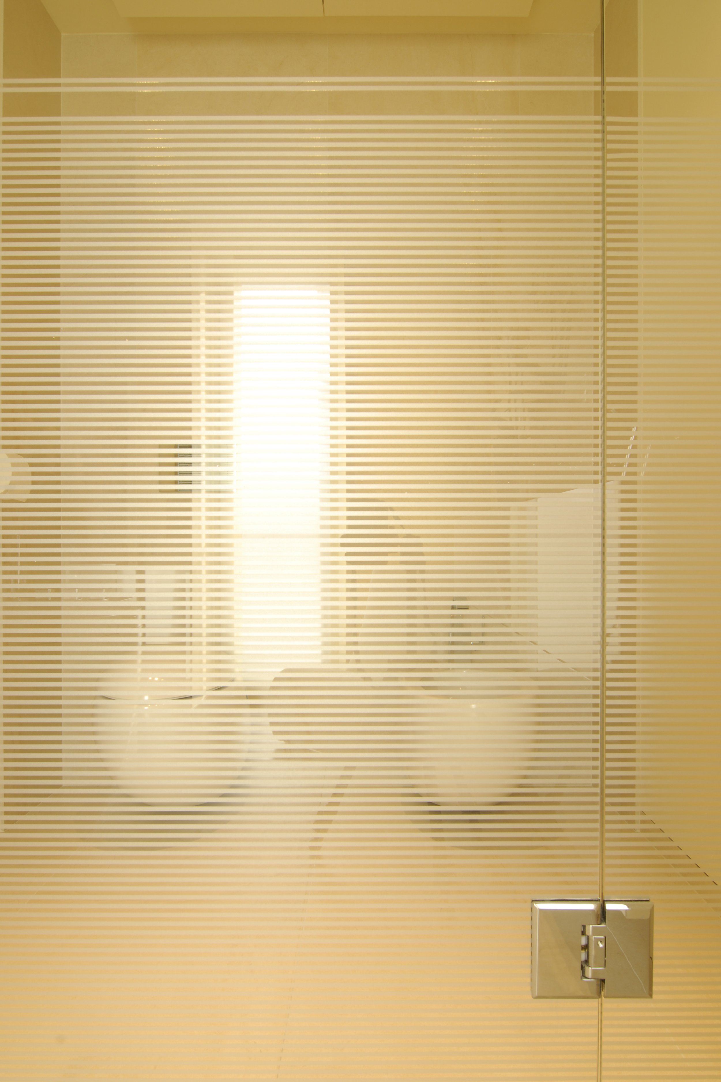 Ein Blick durch eine halbtransparente, gestreifte Tür auf eine moderne Toilette mit weißen WCs und einer silbernen Türsicherung.