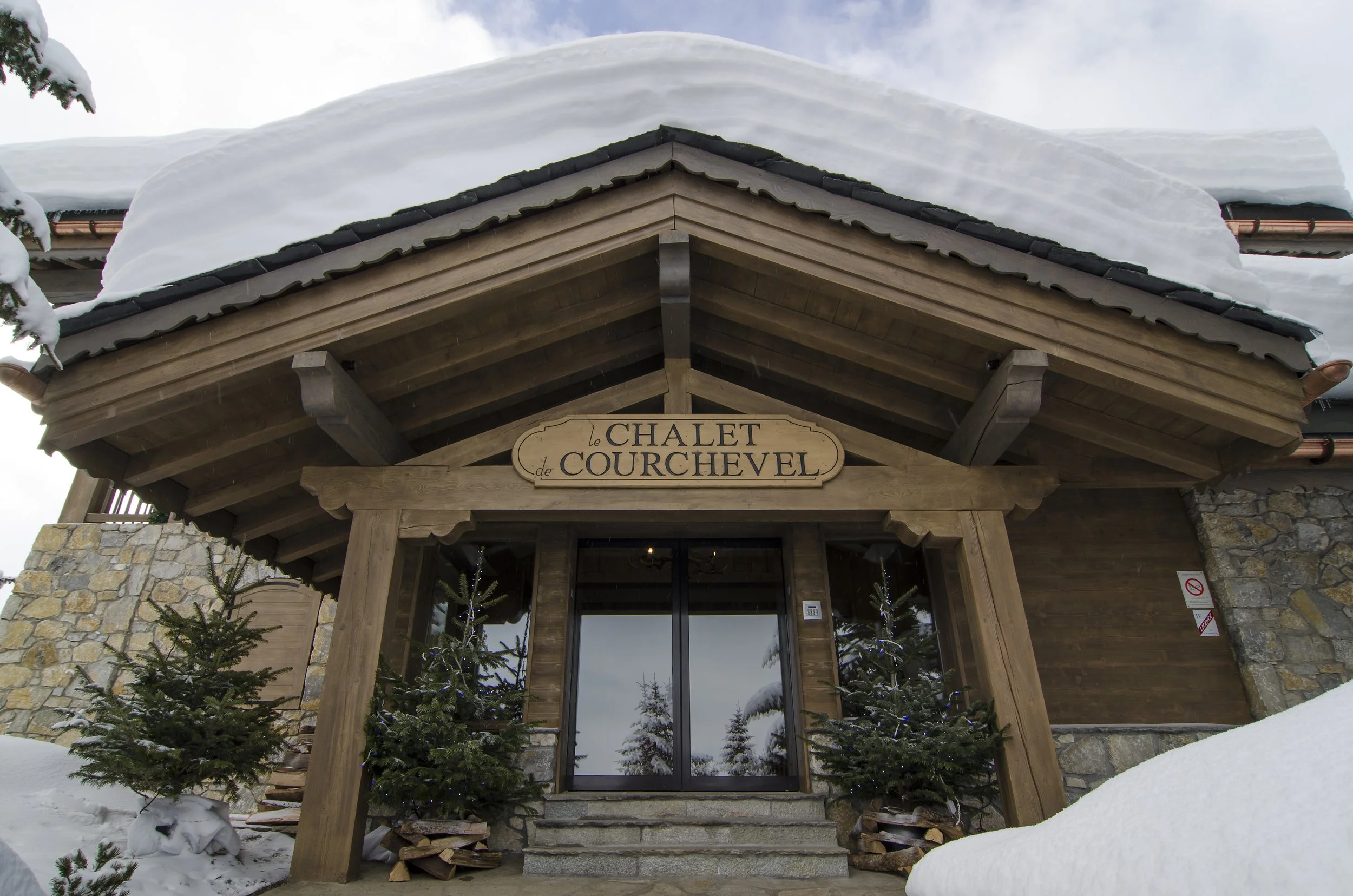 Holzhaus mit Schneedecke auf dem Dach, Weihnachtsbäumen vor dem Eingang und einem Schild mit der Aufschrift 'le CHALET de COURCHEVEL'
