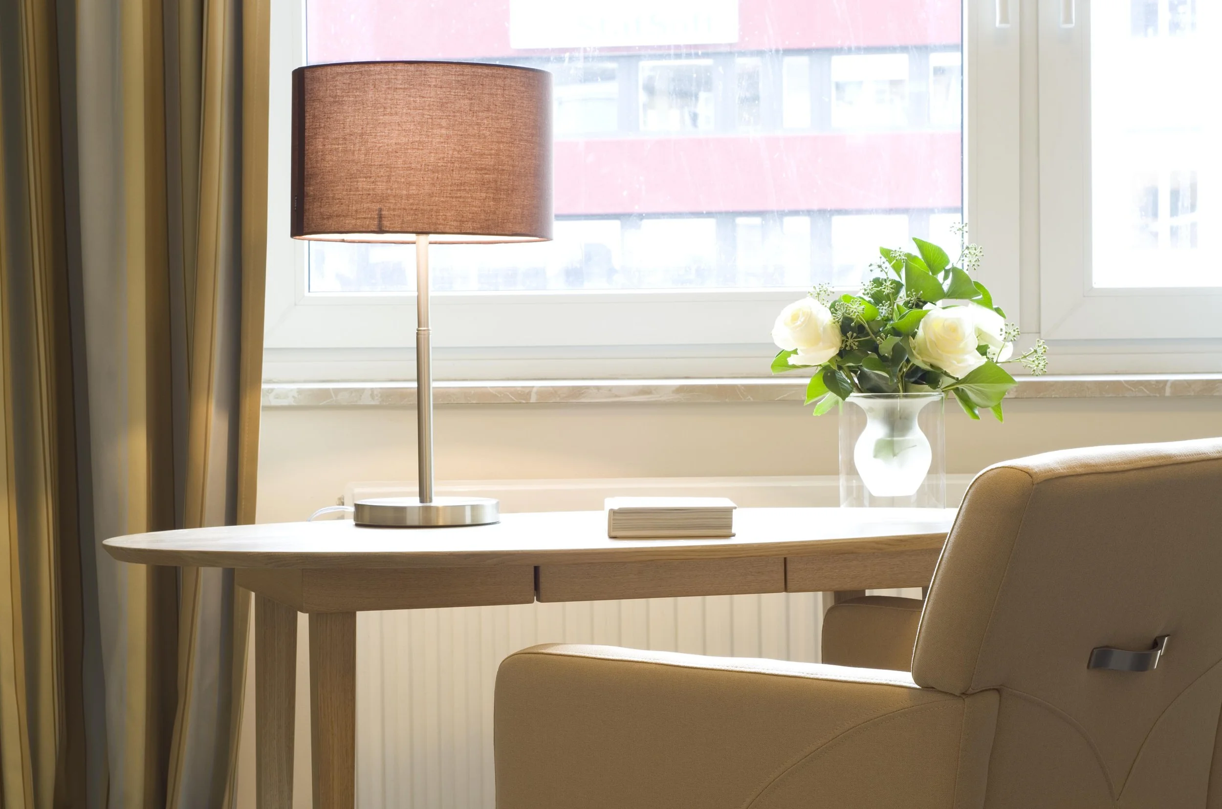 Ein Fenster mit Gardinen, ein Tisch mit einer Stehlampe und einer Vase mit weißen Rosen, im Hintergrund die Außenseite eines Gebäudes.
