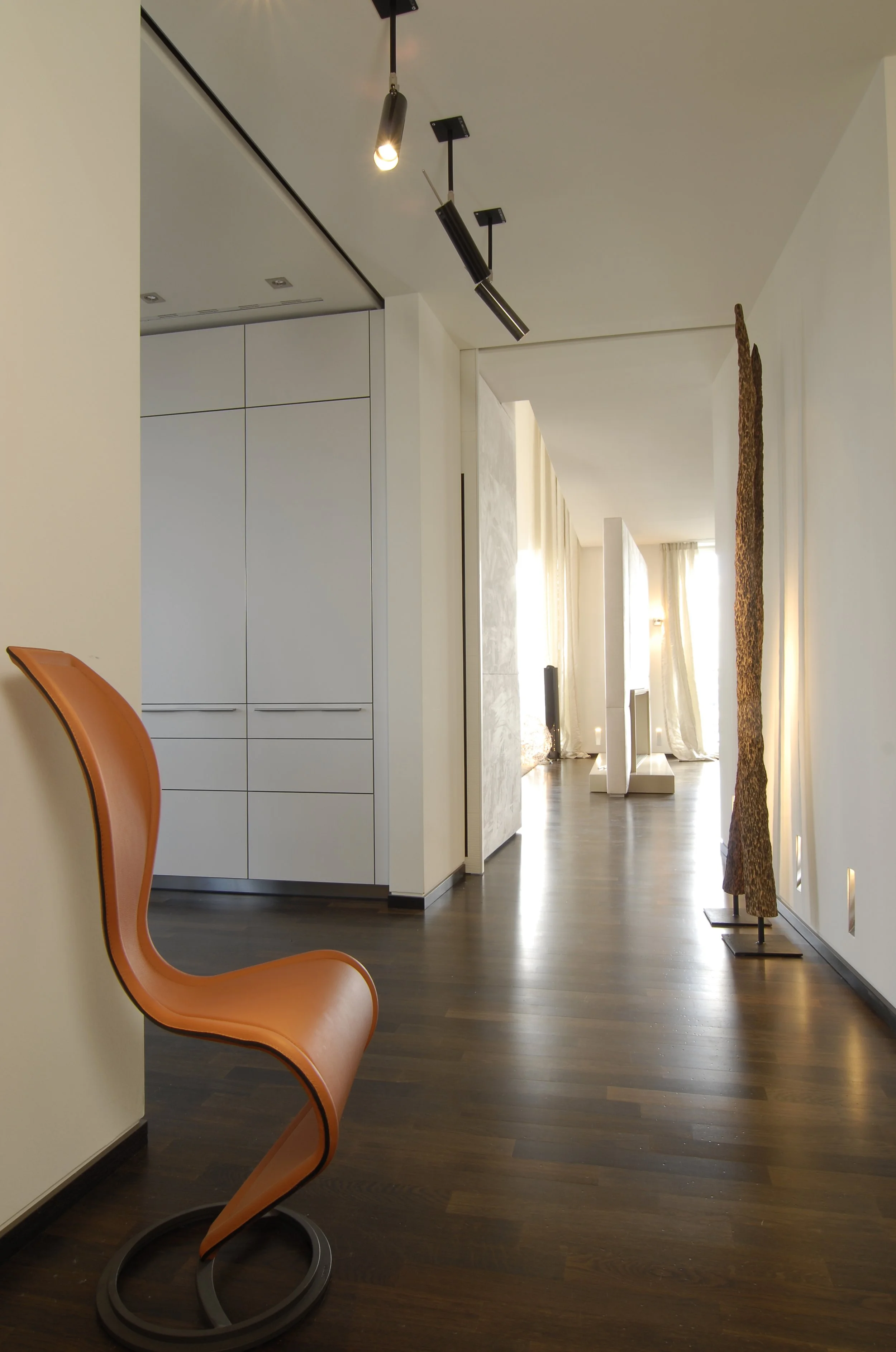 Moderner, heller Flur mit Holzfußboden, orangefarbener Stuhl, Kunstinstallationen an der Wand und Deckenleuchten.