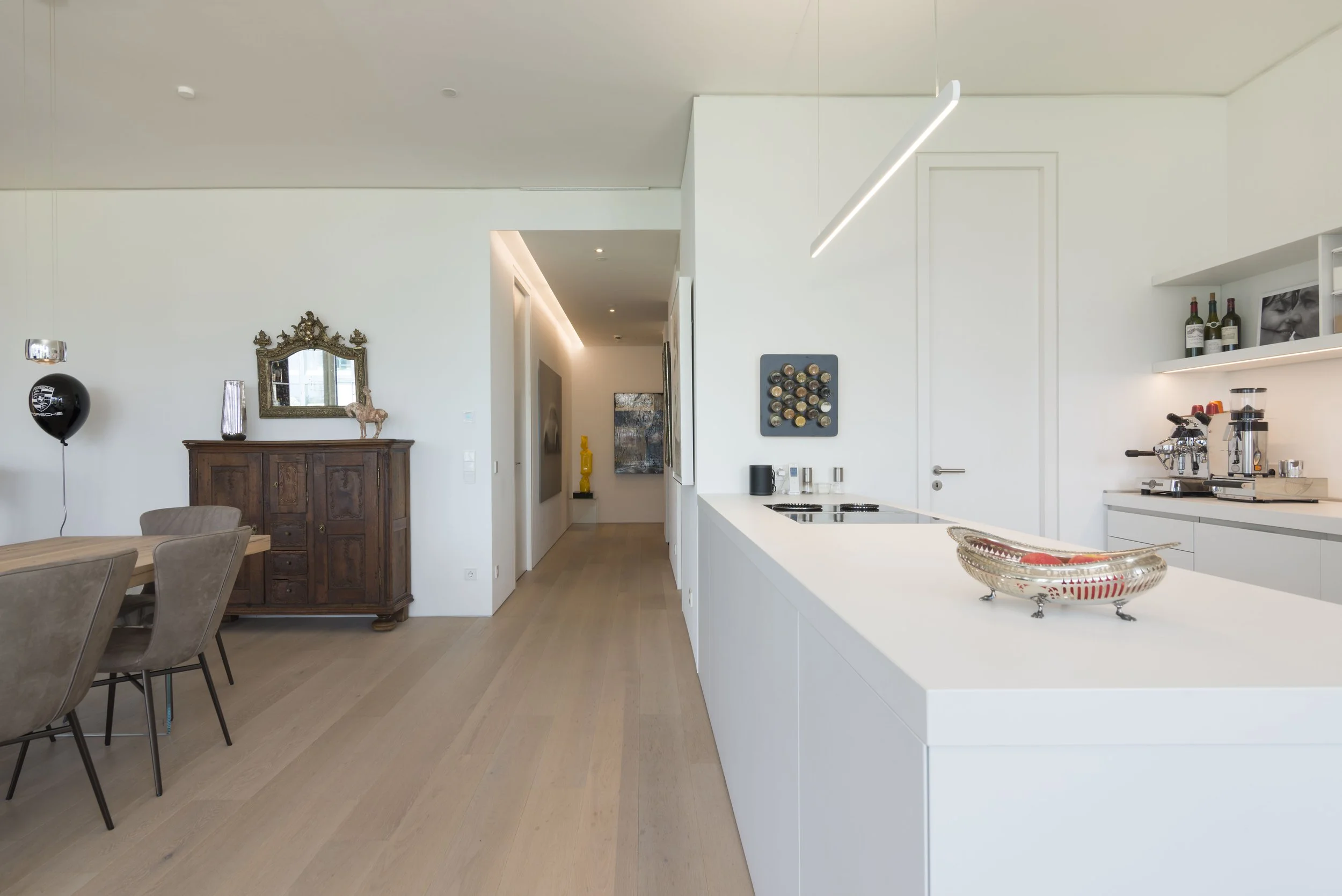 Moderne, offene Küche mit weißem Design, Esstisch mit Stühlen, antiker Holzschrank, Wanddekorationen, Kaffeeautomat, Weinflaschen.
