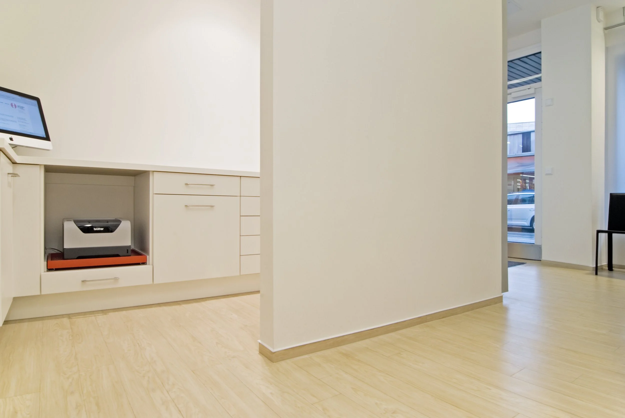 Innenansicht eines modernen, hellen Warteraums mit weißen Wänden, Holzboden und einer Tür, die auf einen Eingangsbereich mit Glasfront führt.