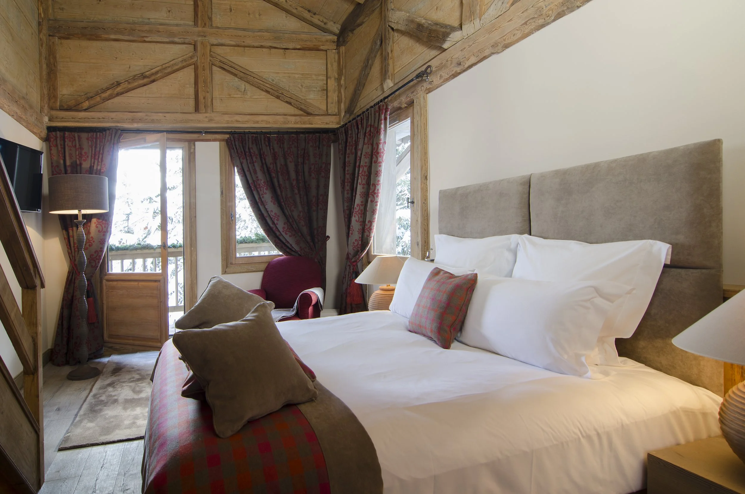 Schlafzimmer in einer Berghütte mit Holzbalken, Bett mit weißen Bettwäsche, Kissen und Tagesdecke, Fenster mit roten Vorhängen, ein Sessel und Nachttische mit Lampen.