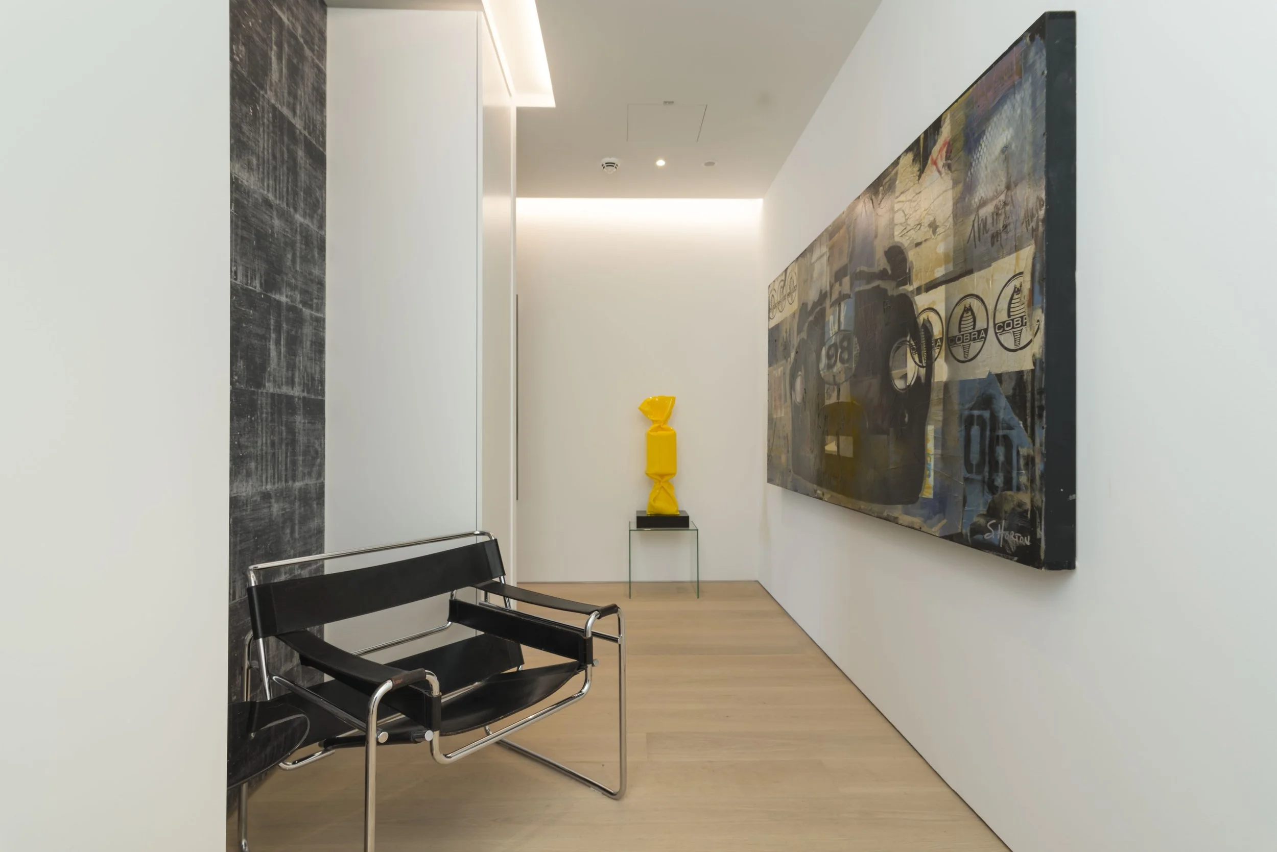 Ein moderner Kunstgalerie-Flur mit schwarzem Sitzbank, schwarzer Wand, weißen Wänden, großem abstraktem Gemälde an der rechten Wand und skulpturartiger Skulptur auf einem kleinen Glasständer in der Mitte des Flurs.