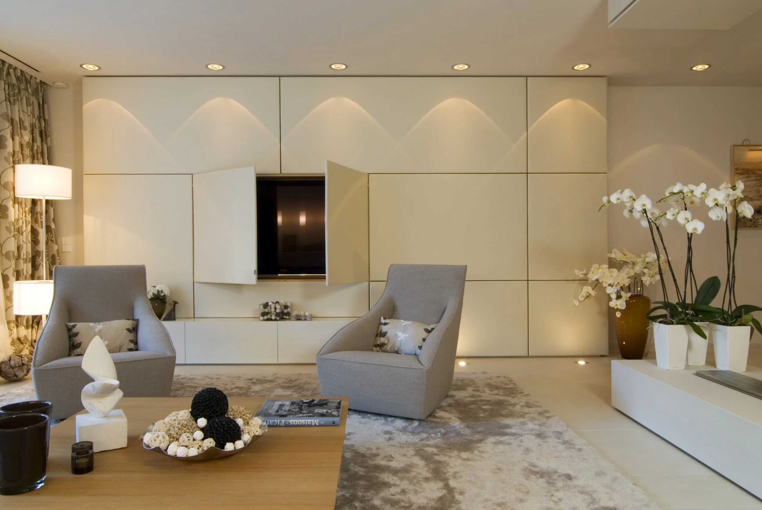 Moderne Wohnzimmer mit weißen Wänden, zwei grauen Sessel, einem Holztisch mit Dekorationen, einem großen weißen Schrank, Orchideen in weißen Töpfen und indirektem Beleuchtung.