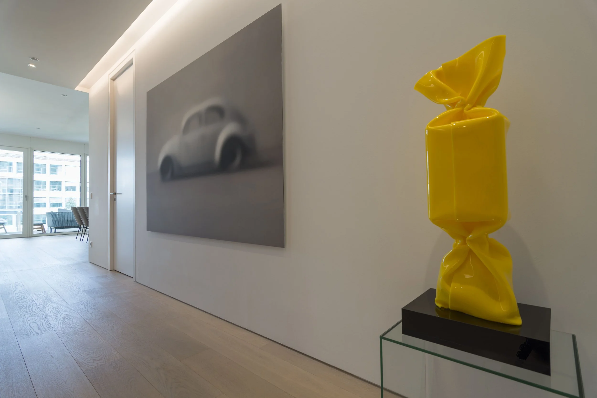 Moderne Ausstellung mit einer gelben Skulptur aus verschmolzenem Kunststoff auf einem schwarzen Sockel, daneben ein bläulich-graues Wandbild eines verwaschenen Autos in einem hellen Ausstellungsraum
