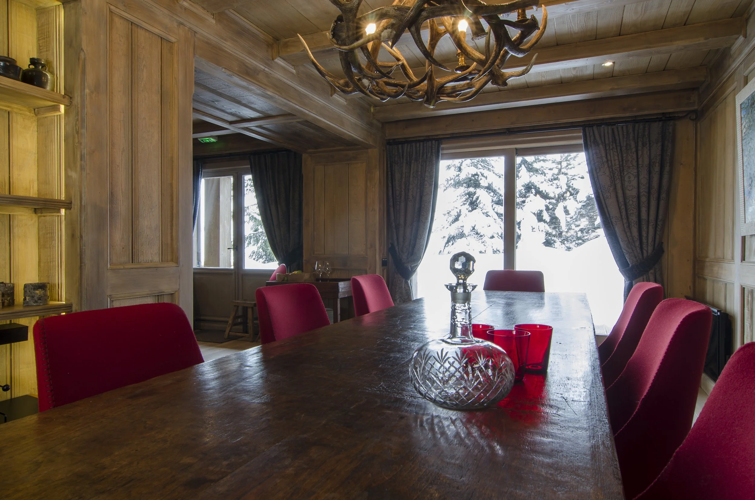 Ein rustikaler Holztisch mit roten Stühlen in einem gemütlichen, holzverkleideten Esszimmer. Im Hintergrund sind Fenster mit dunklen Vorhängen und ein Schneelandschaft im Winter zu sehen.