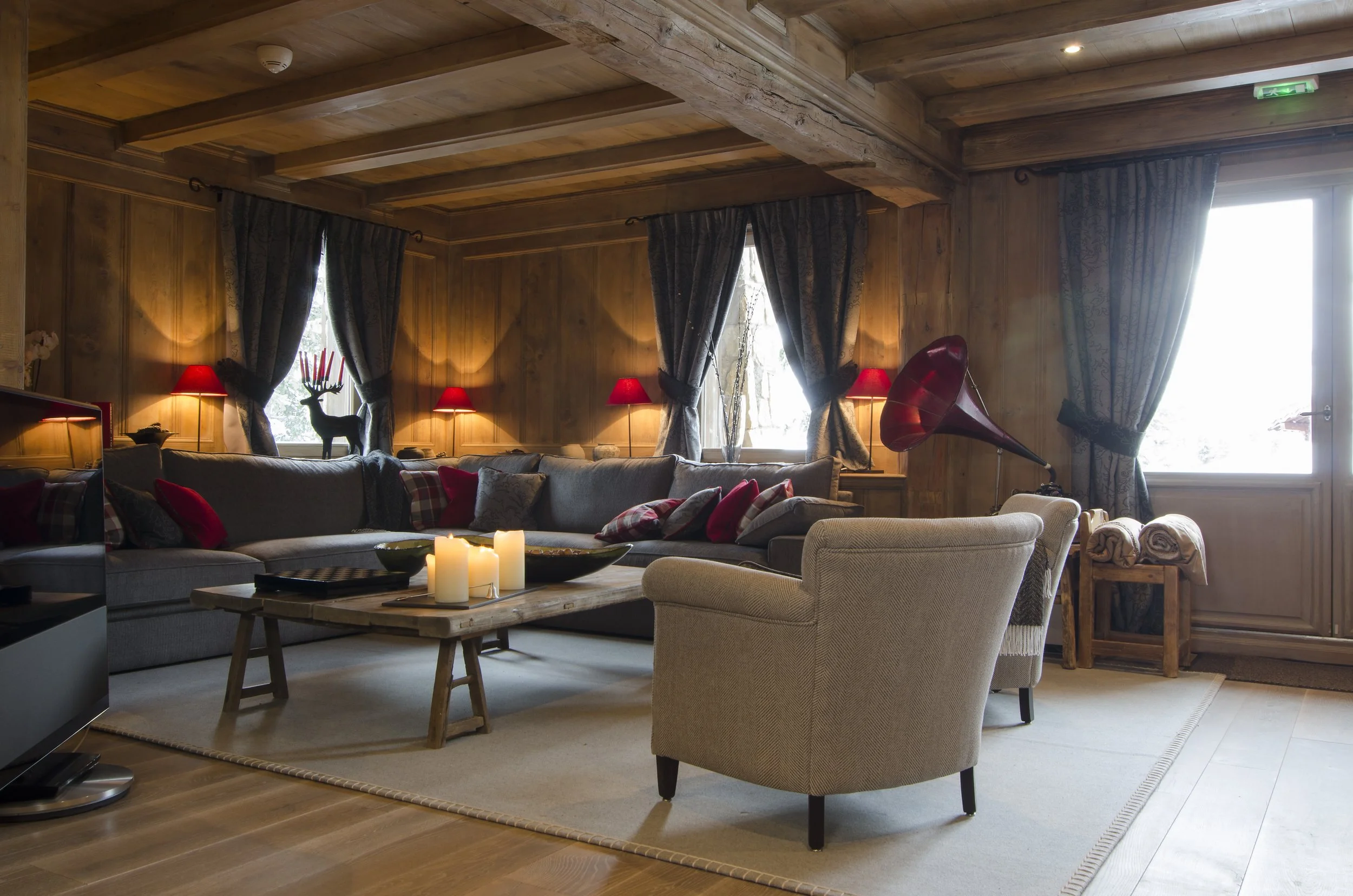 Gemütliches Wohnzimmer mit Holzwänden und Decke, großer grauer Sofa mit roten und karierten Kissen, ein beiger Sessel, Couchtisch mit Kerzen, Vorhänge und Fenster, alte Grammophon auf einem Tisch, dämmerndes Licht.