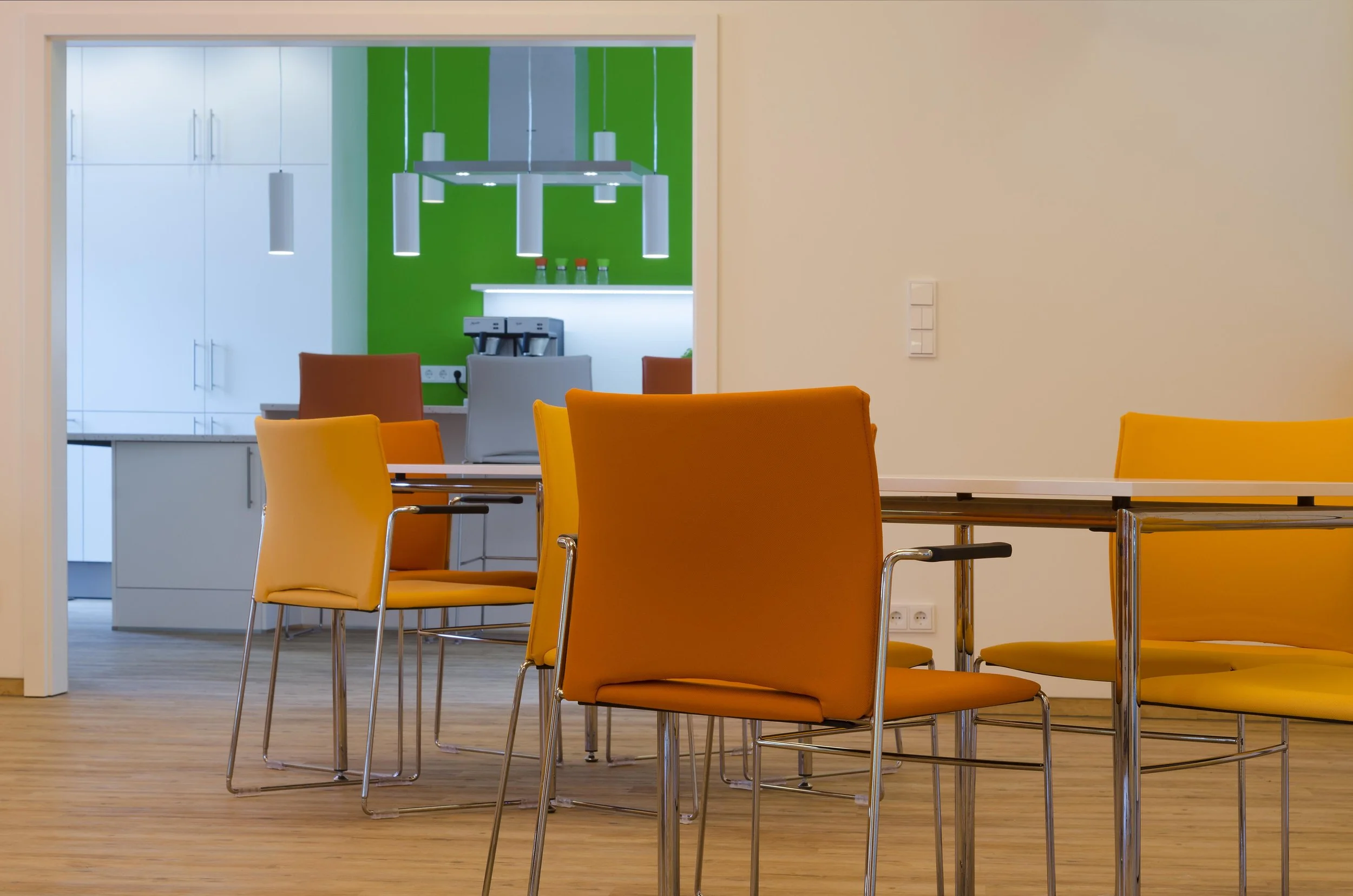 Ein modernes Esszimmer mit orangefarbenen Stühlen um einen Tisch, im Hintergrund eine Küche mit weißen Schränken und einer Kaffeemaschine, ein Sichtfenster führt in die Küche, die Wand ist beige.
