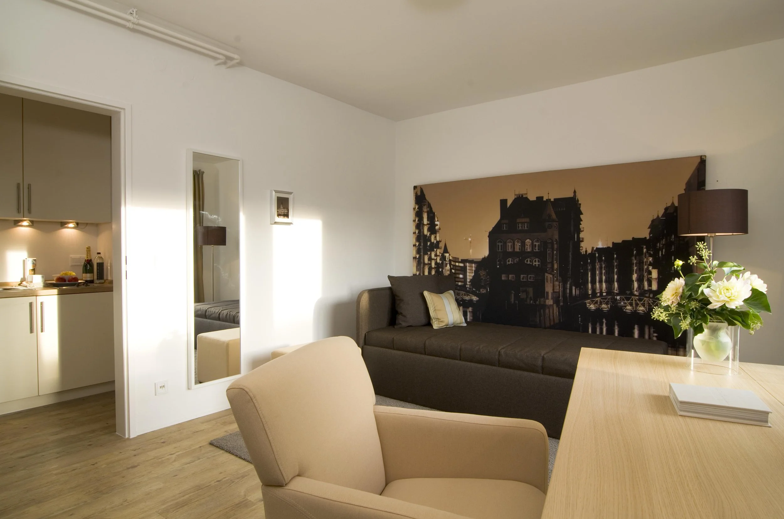 Wohnzimmer mit Bett, Sessel, Blumen, Wandbild und Blick in die offenen Küche im Hintergrund.