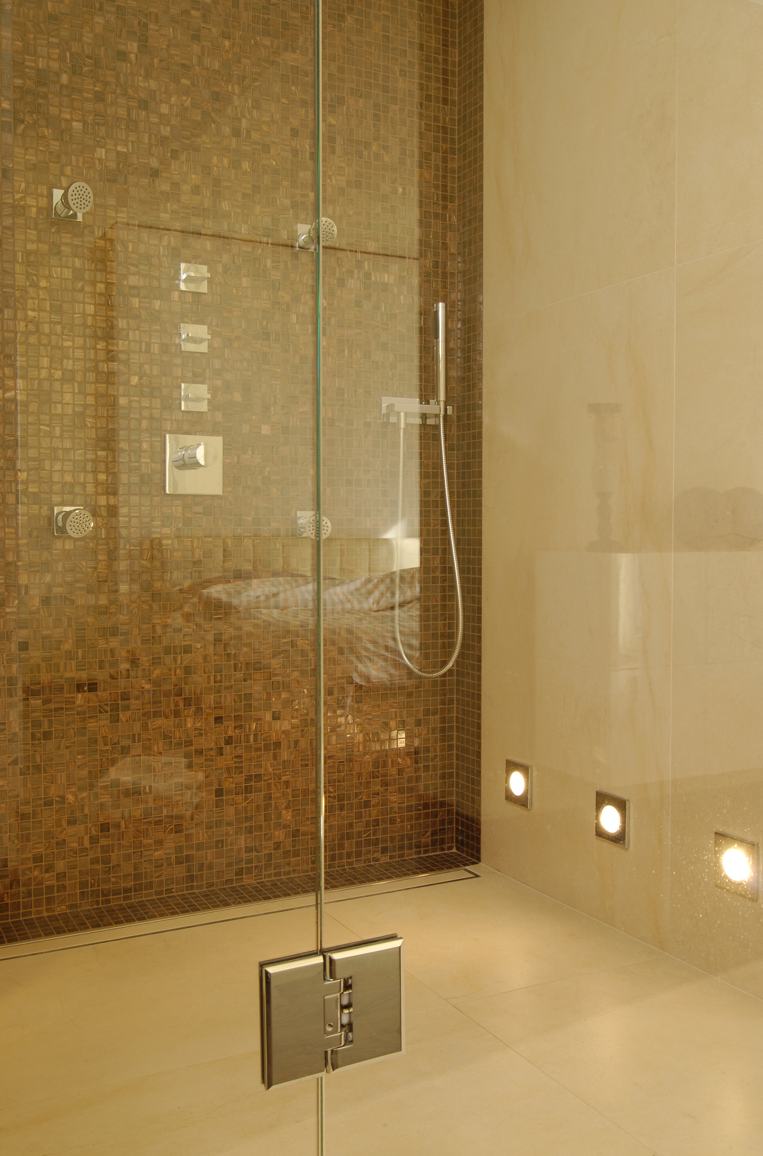Moderner Duschbereich mit Brausekopf, Handbrause und unterschiedlichen Wasserreglern, hinter einer Glaswand in einem Badezimmer mit beige-hellbraunen Fliesen.