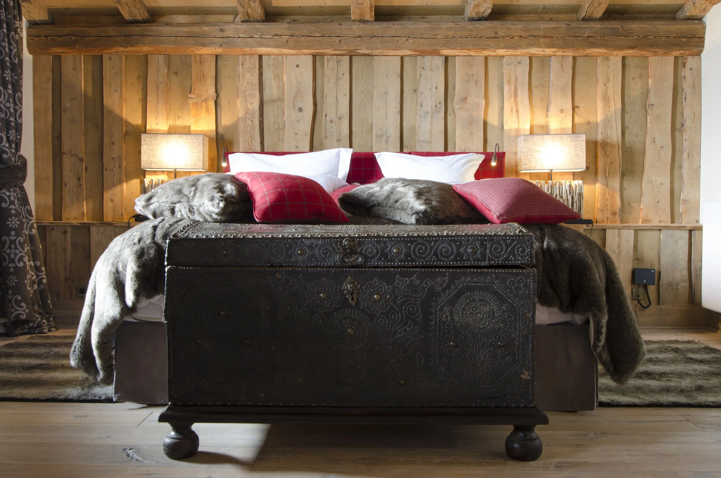 Ein gemütliches Schlafzimmer mit Holzverkleidung, Bettruhe und dekorativen Kissen sowie Decken aus Fell. Zwei Nachttische mit Lampen sind auf beiden Seiten des Bettes. Es gibt einen Teppich auf dem Holzboden.