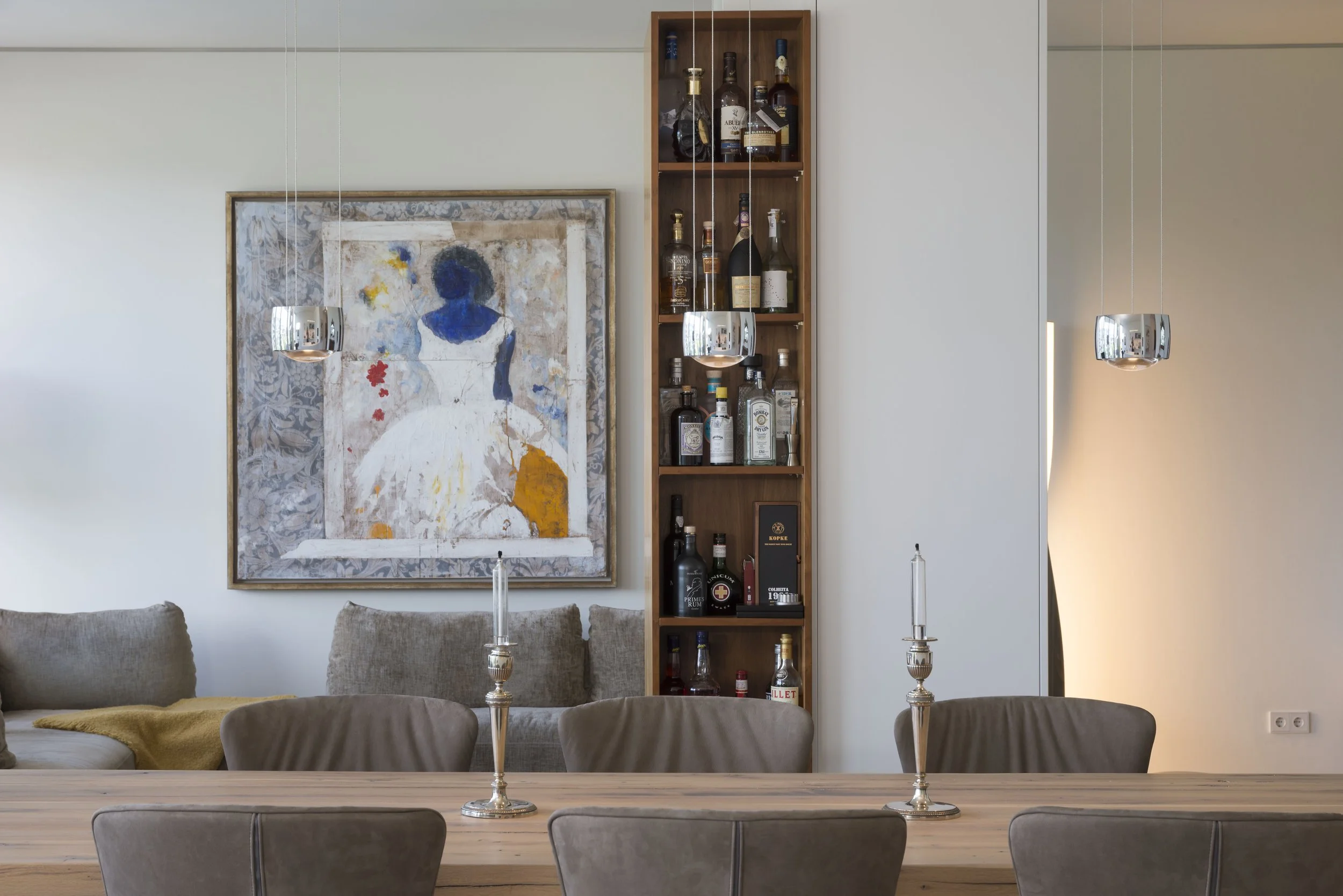 Ein Esszimmertisch mit sechs Stühlen, zwei silberne Kerzenleuchter, im Hintergrund ein Sofa, eine große Wandkunst mit abstrahierter Frau, ein offener Regal für Spirituosen, weiße Wände, moderner Wohnstil.