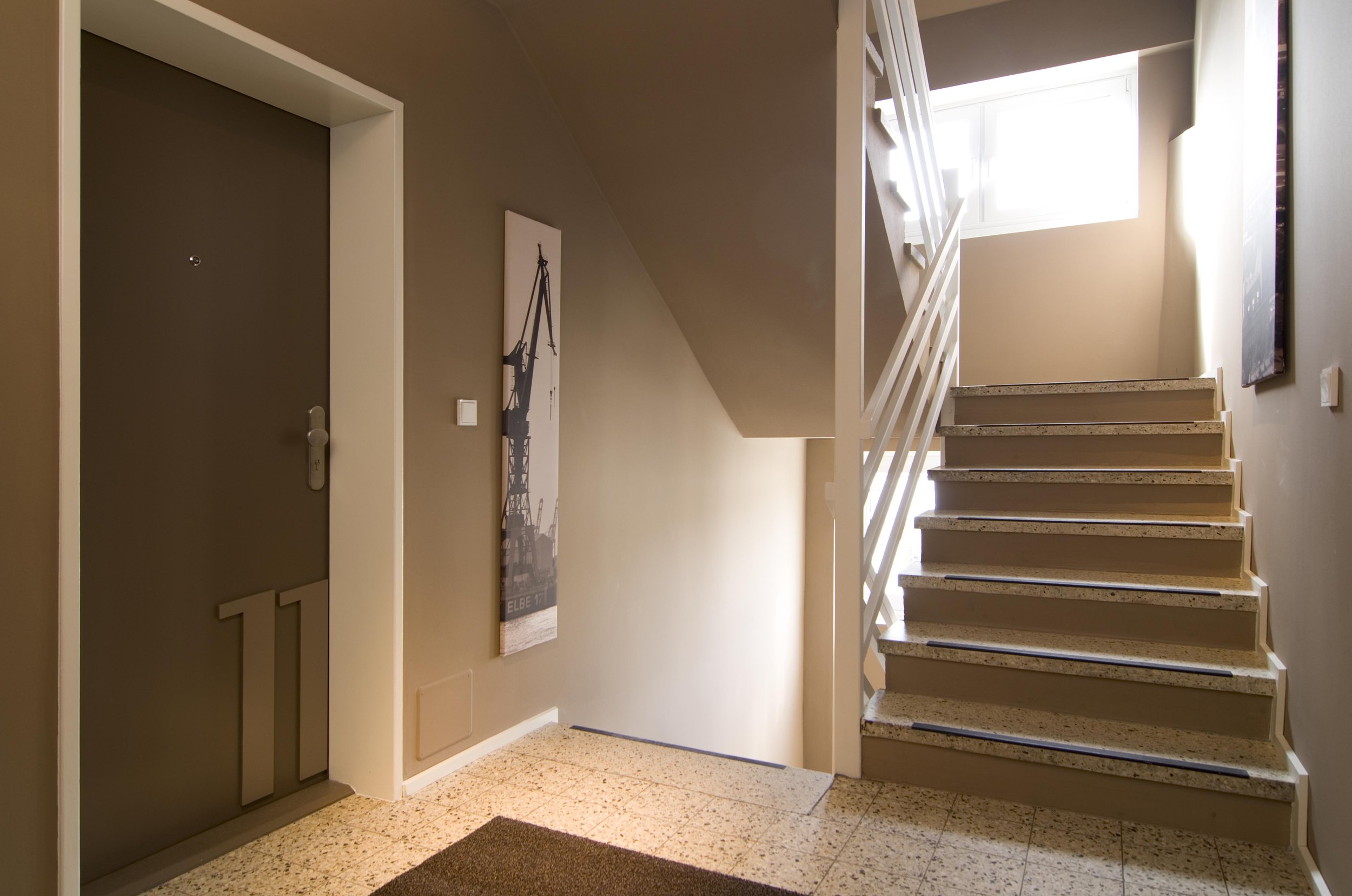 Innenaufnahme eines Flurs mit Treppenhaus, graue Wand, brauner Fußboden, ein grauer Tür mit der Nummer 11, ein Wandbild eines Kranes, und ein Fenster oberhalb der Treppe, das Licht strömt herein.