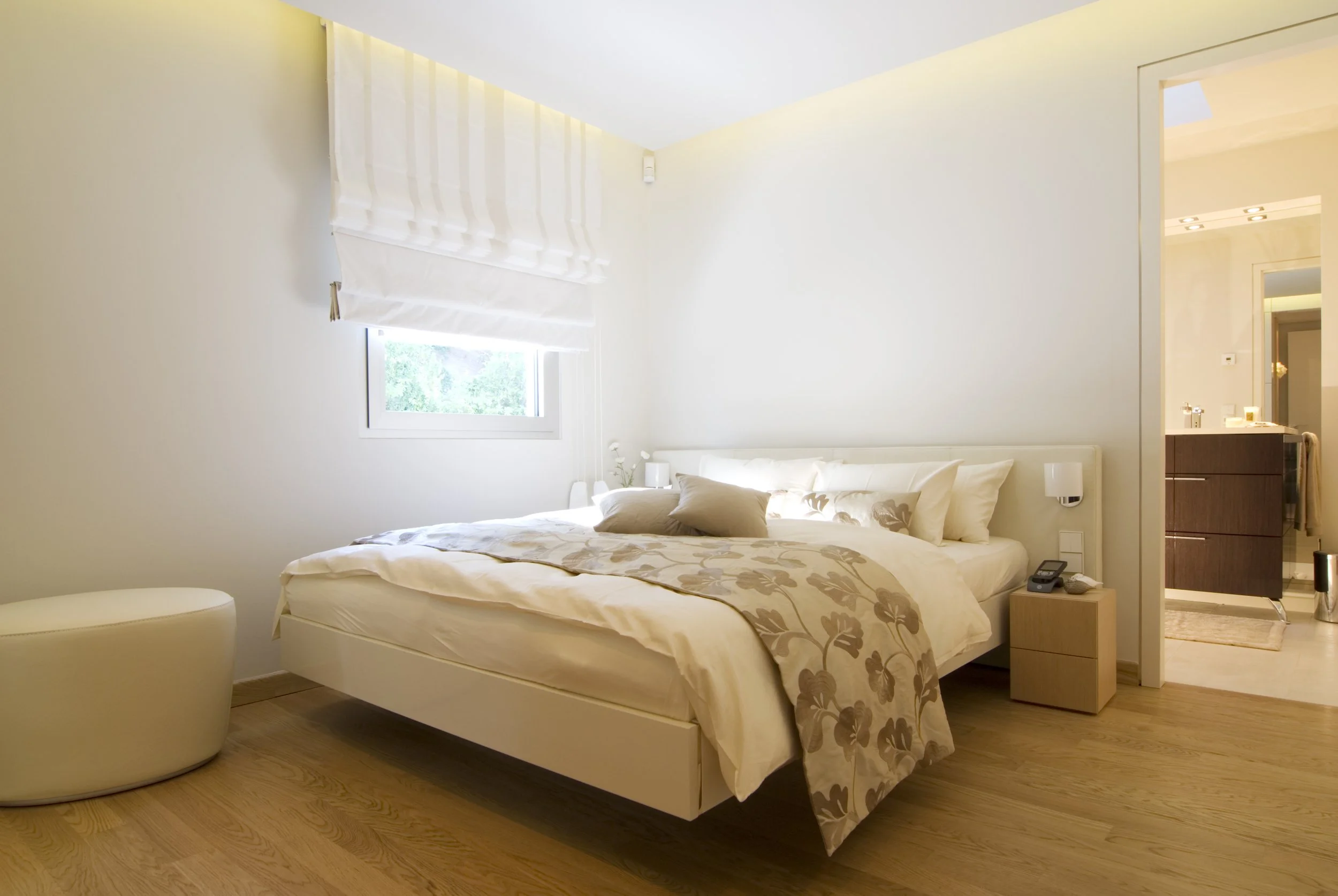 Ein modernes Schlafzimmer mit einem großen weißen Bett, beige und weißen Bettwäsche, einem kleinen Nachttisch mit Telefon und Licht, einem kleinen weißen Sessel, einem Fenster mit weißer Rolllade und Blick nach draußen, und einem offenen Badezimmer m