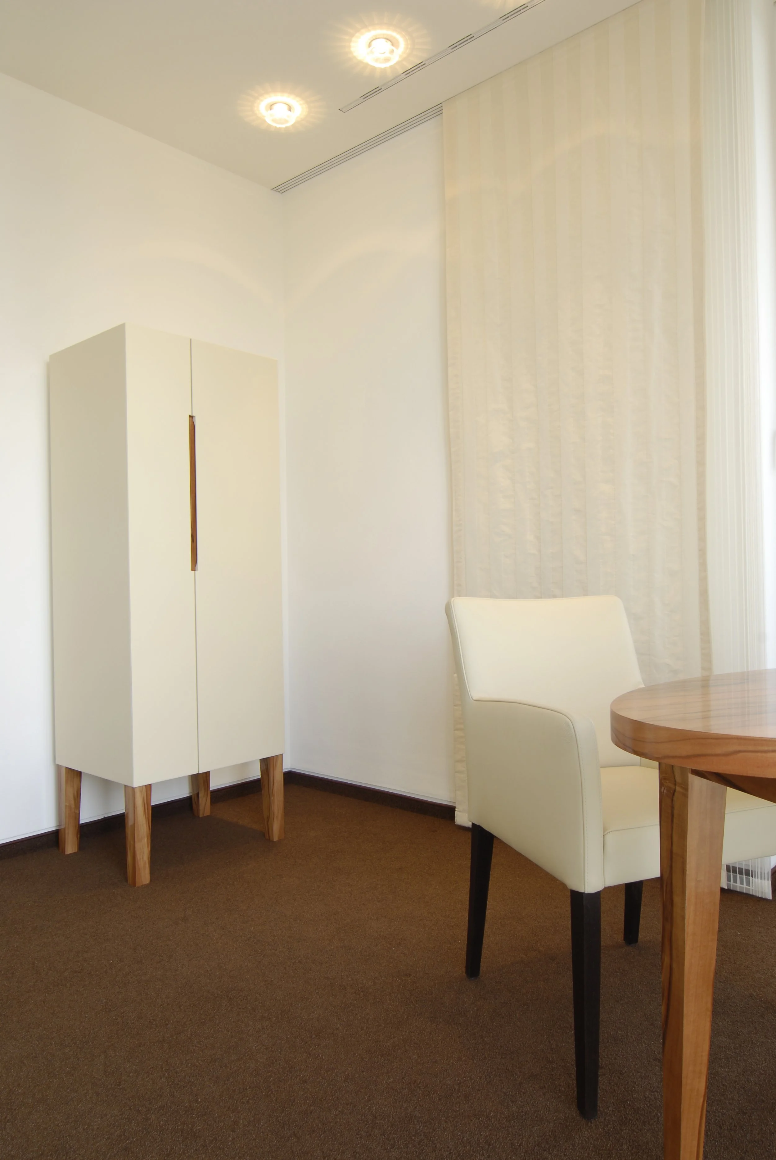 Ein weißes Möbelstück auf Holzkufen, ein weißer Stuhl und ein runder Holztisch vor weißen Wänden und Gardinen, Deckenleuchte an der Decke.