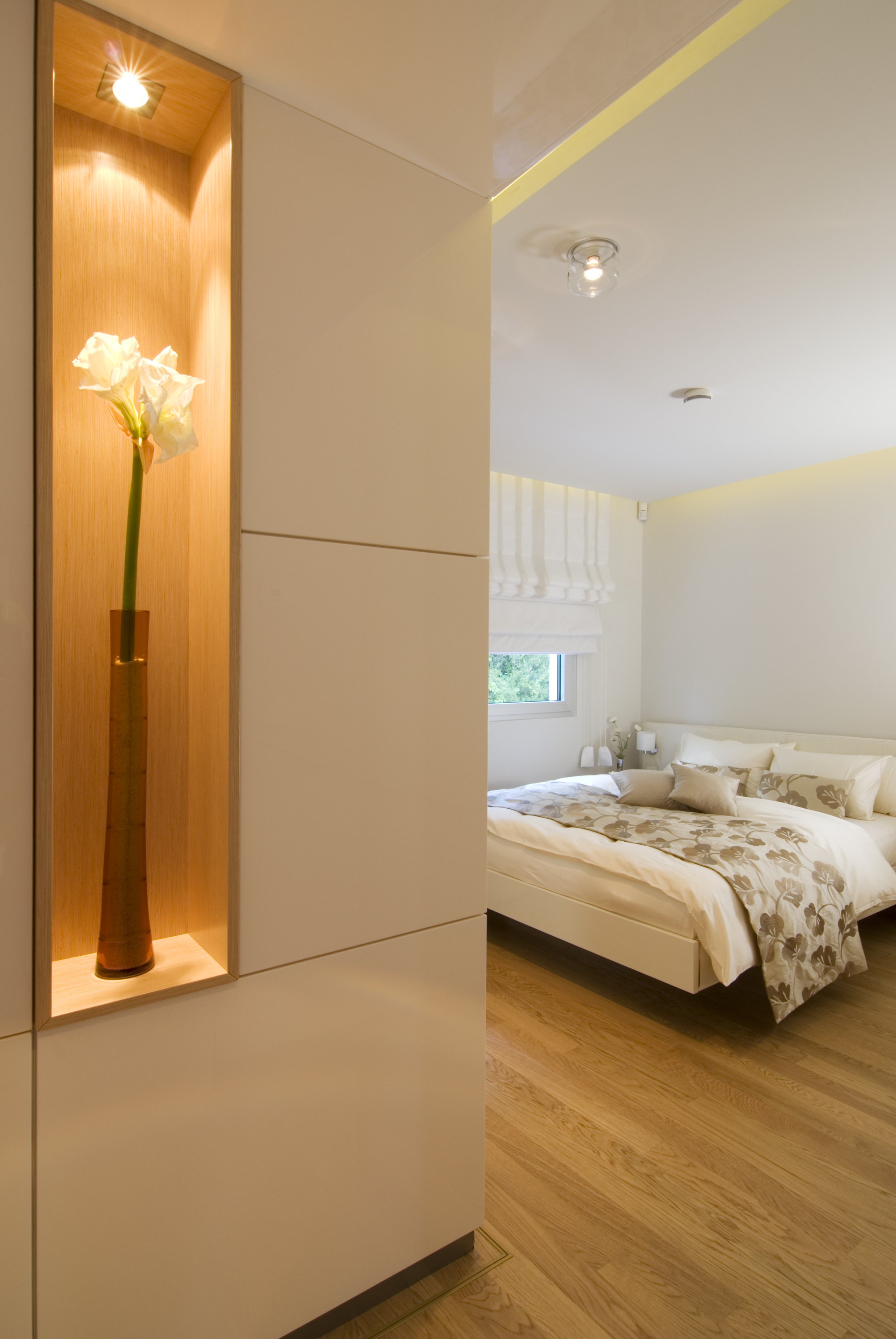 Modernes Schlafzimmer mit weißem Bett, dekorativen Kissen, Holzfußboden und einer Fenster mit weißen Vorhängen. Im Vordergrund eine gepflanzte Vase mit weißen Blumen in einer eleganten Nische an der Wand.
