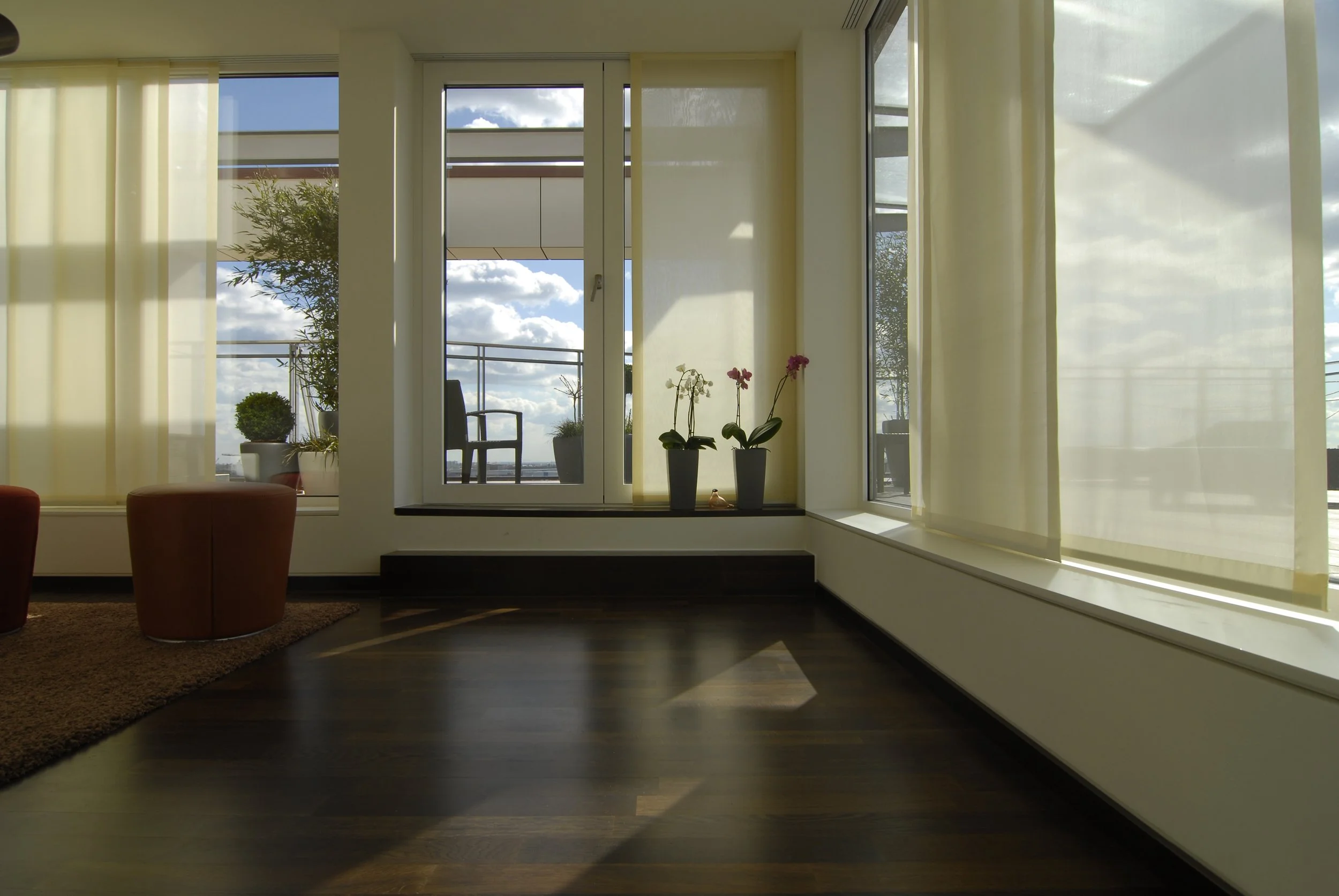 Ein heller Wohnraum mit großen Fenstern und Blick auf einen Balkon mit Stuhl und Pflanzen. Im Vordergrund befinden sich zwei Töpfe mit Blumen, Sonnenlicht durch die Fenster. 