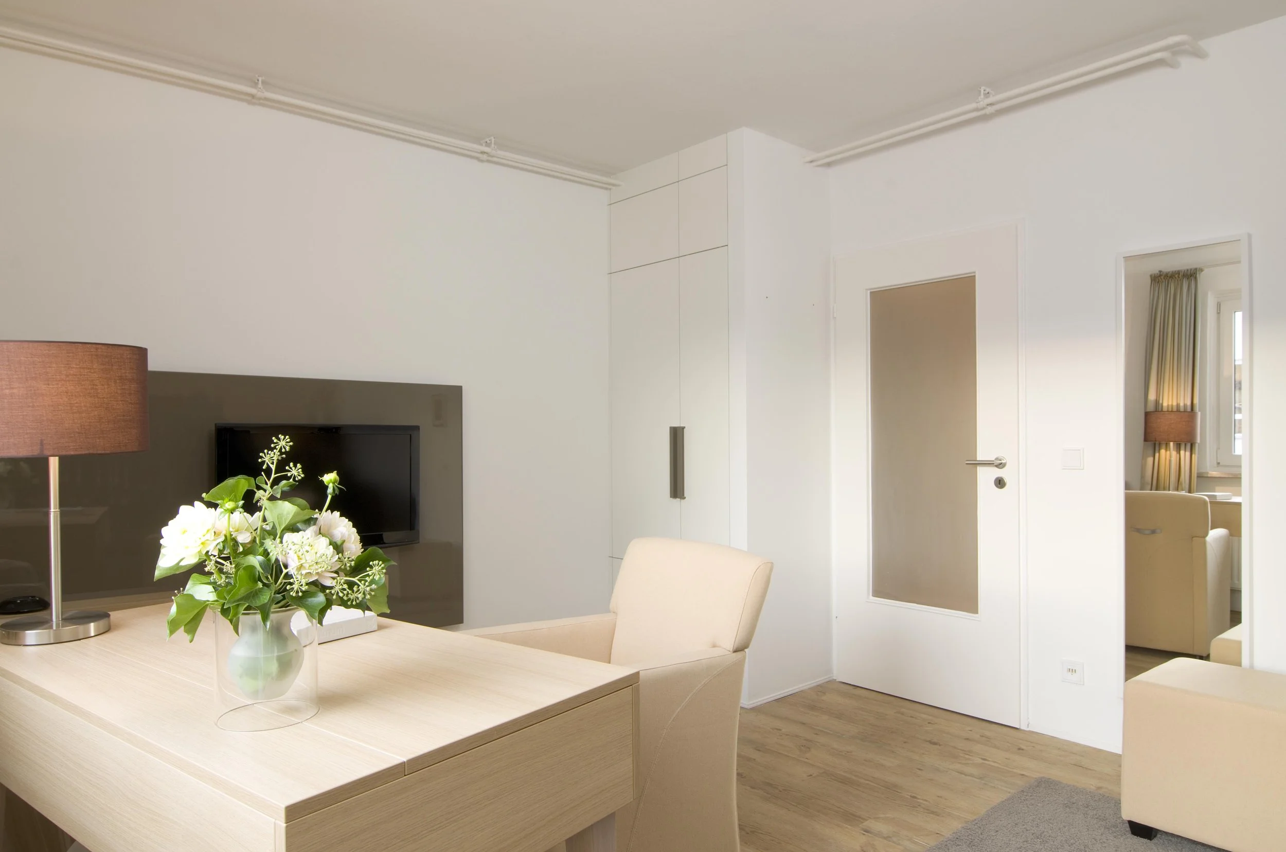 Innenansicht eines hellen, modernen Wohnzimmers mit hellen Möbeln, einem Blumentopf mit weißen Blumen auf einem Tisch, einem Fernseher an der Wand, einer weißen Tür und einem Fenster mit Vorhängen.