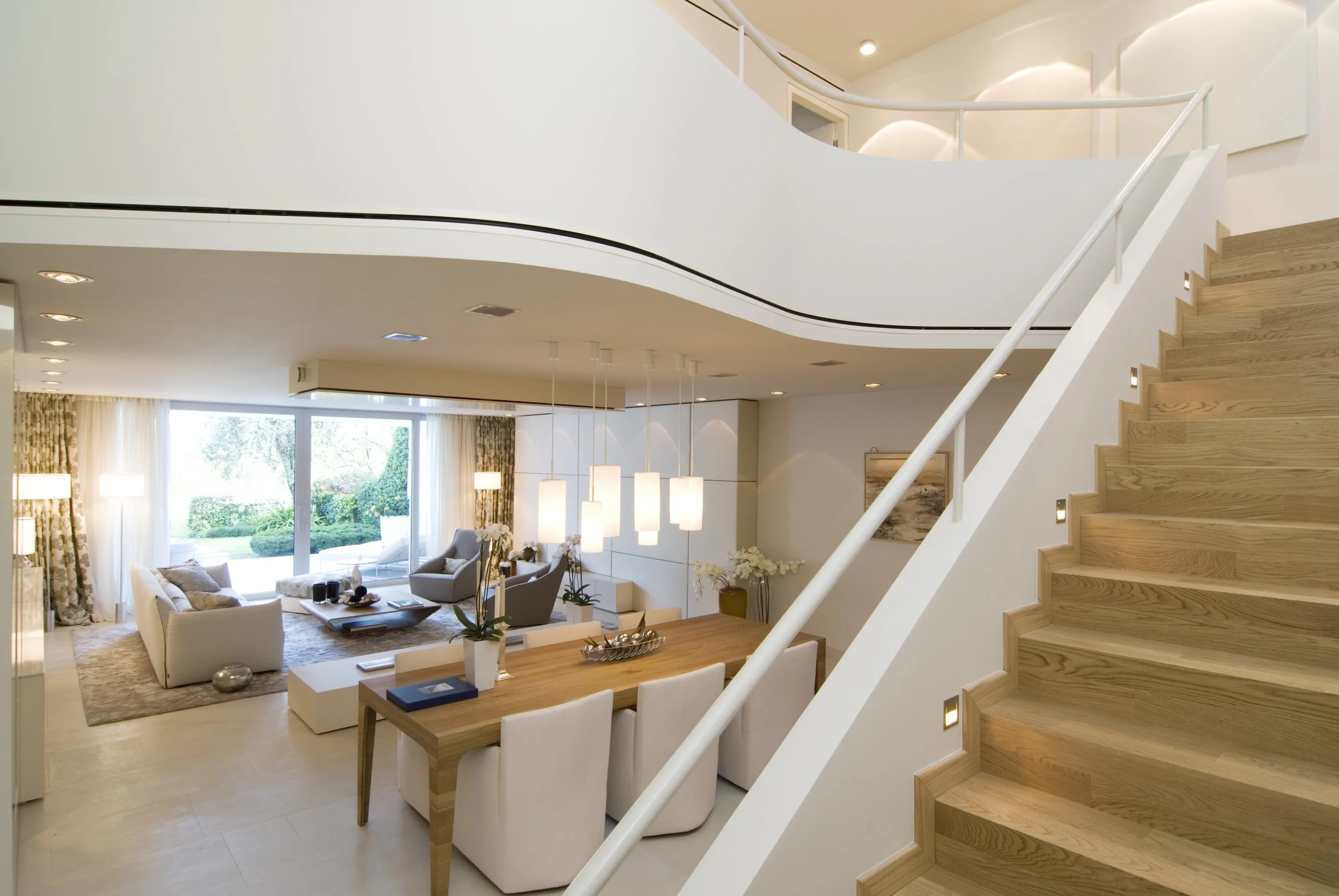 Modernes Wohnzimmer mit offener Treppe, großen Fenstern, gemütlicher Sitzgruppe, Essbereich mit Hängeleuchten, und Blick auf Garten durch Fenster