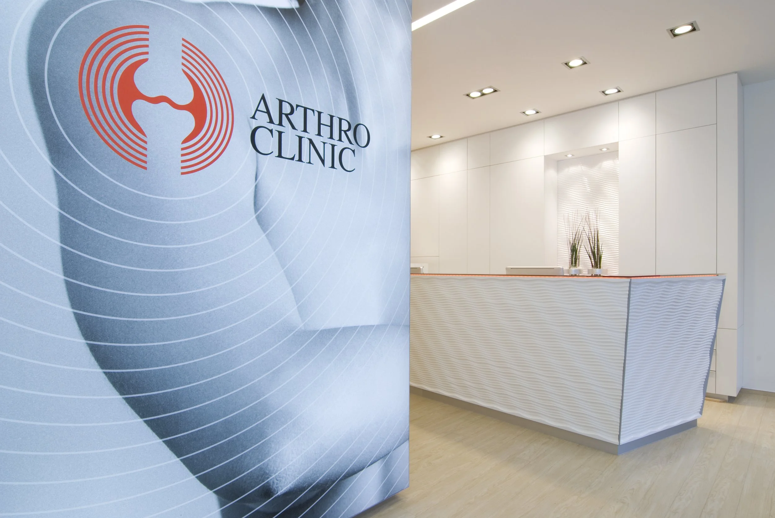 Empfangstresen im Arthro Clinic mit einem großen Logo an der Wand, weißes Design, Pflanzen auf dem Tresen und helle Beleuchtung.