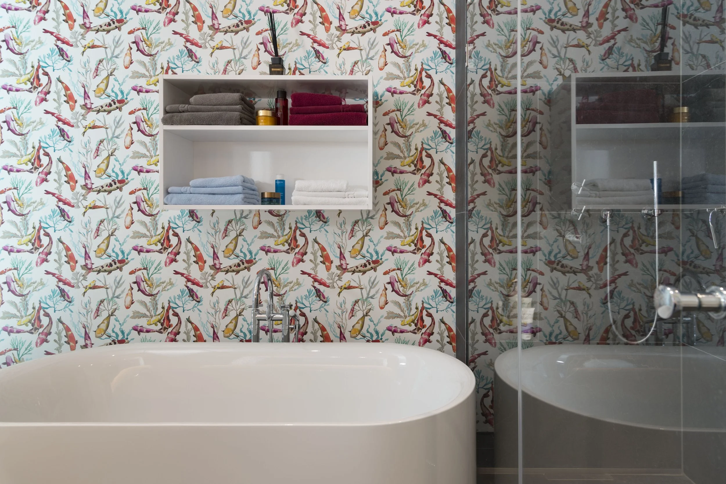 Badezimmer mit buntem Fischtapeten, freistehende Badewanne, Wandregal mit Handtüchern und Pflegeprodukten, Dusche mit Glastrennwand.