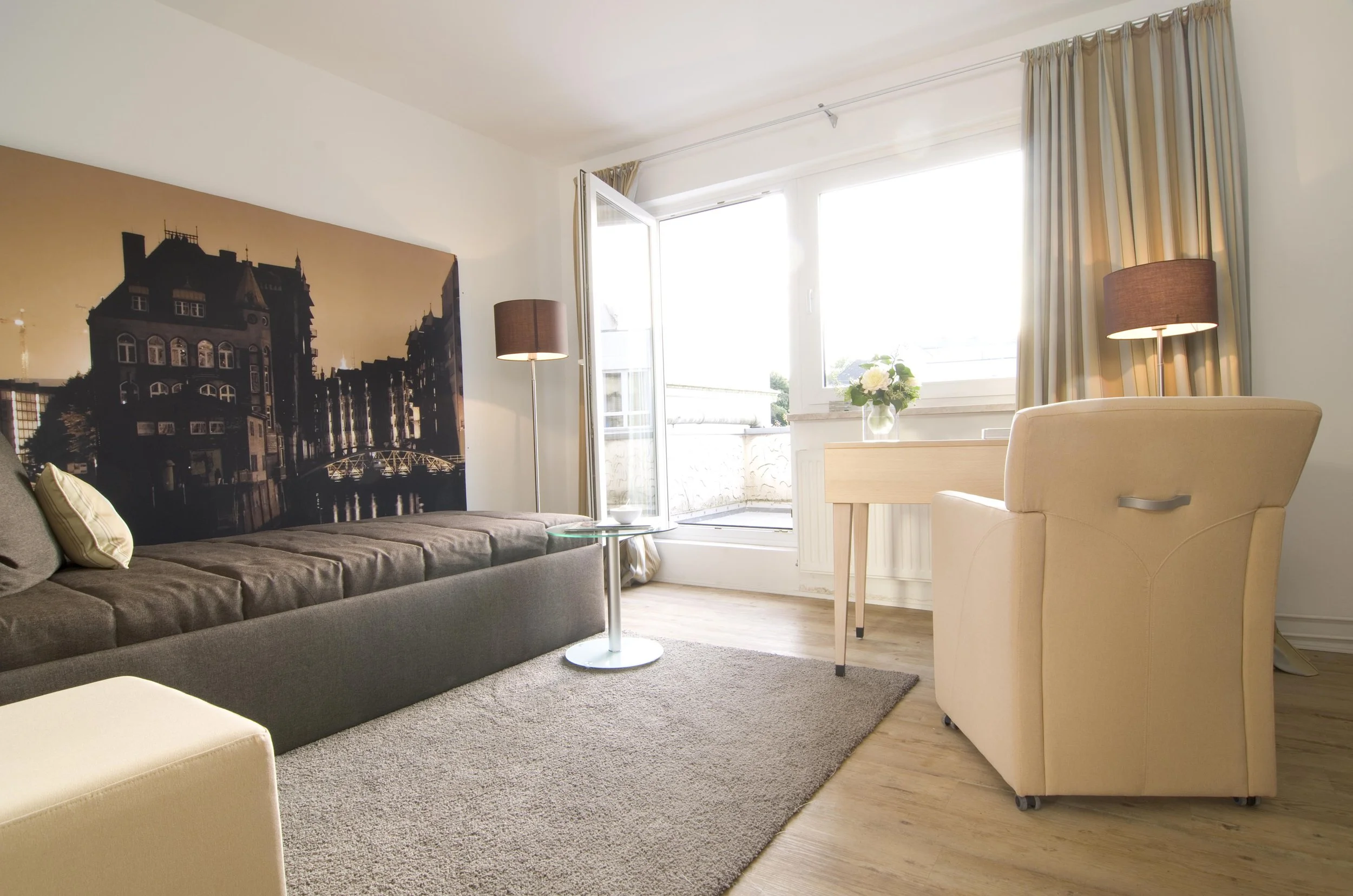 Modernes Wohnzimmer mit Sofa, beigen Sessel, Couchtisch, Stehlampen, großem Fenster mit Blick auf einen Balkon, Vase mit Blumen auf dem Tisch, Holzboden.