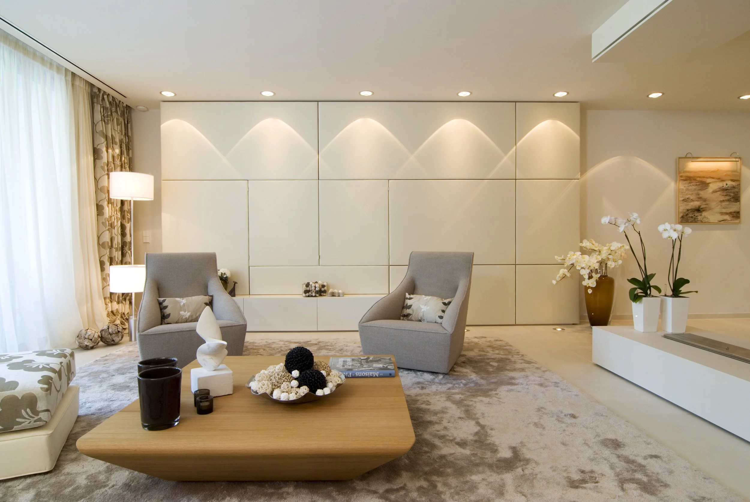 Modernes Wohnzimmer mit grauen Sesseln, Holz-Couchtisch, weißen Wänden und Dekorationen wie Kerzen, Vasen mit weißen Blumen und Bilder an der Wand.