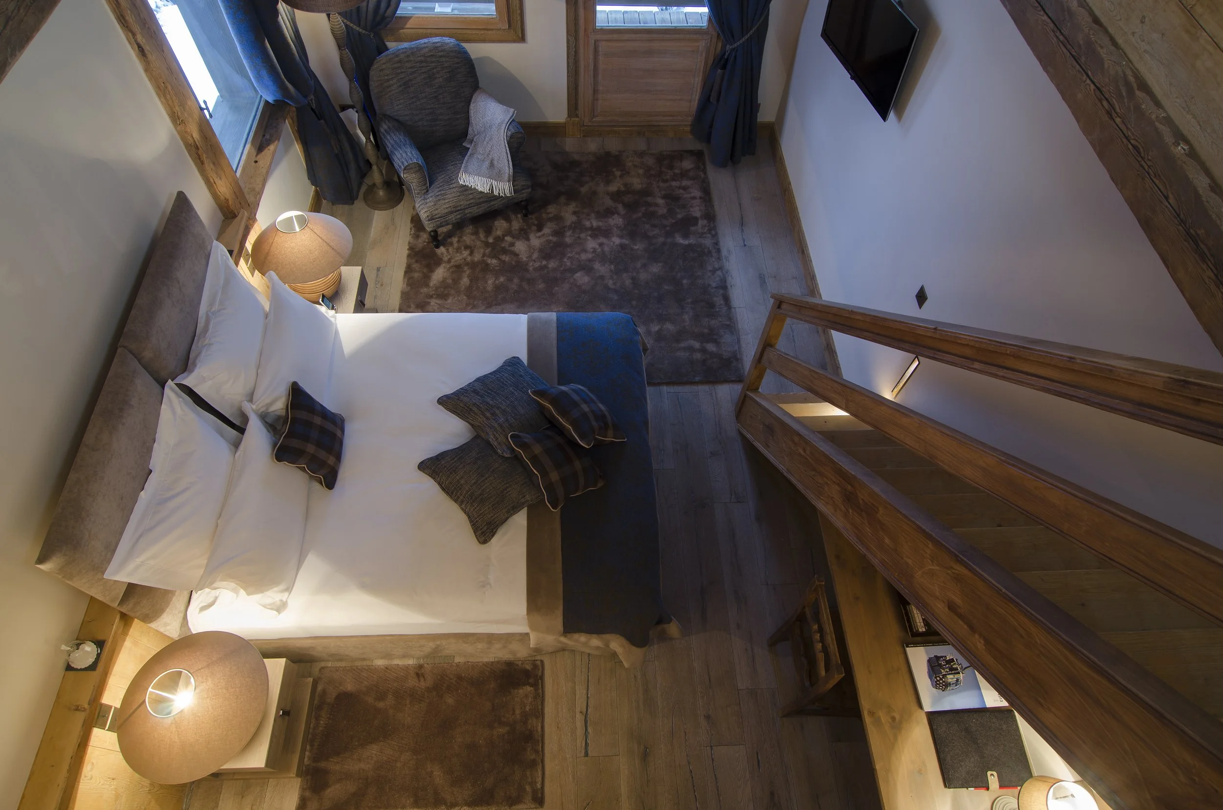 Ansicht eines gemütlichen Schlafzimmers mit Doppelbett, Nachttischen mit Lampen, einem Sessel, einem Fernseher an der Wand, Holzboden, Teppichen und einem Fenster mit Vorhängen.