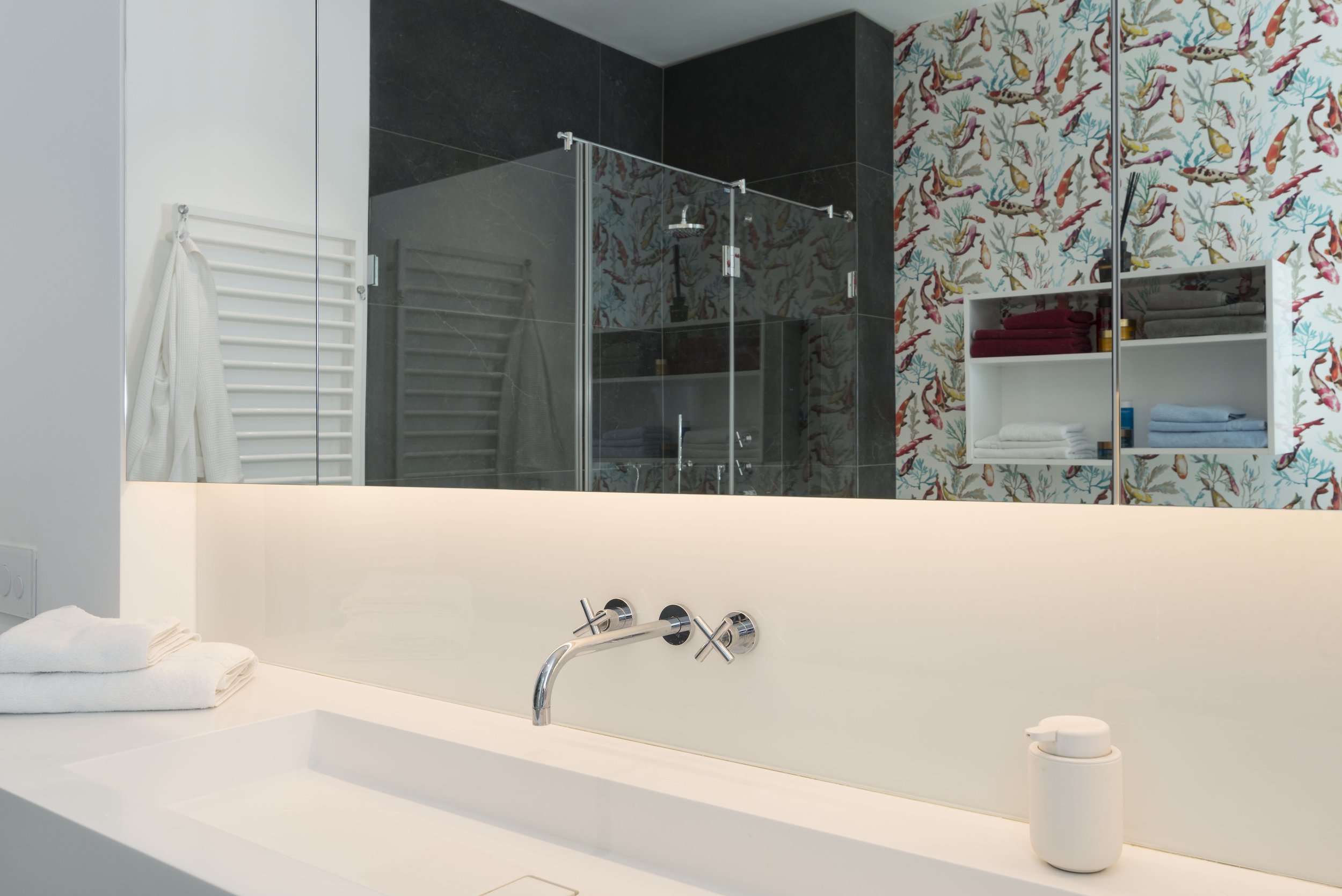 Moderner Badezimmerblick auf eine weiße Waschtischplatte mit Handtüchern, einem weißen Behälter, Spiegel, Badewanne, Dusche im Hintergrund mit dunklen Fliesen und bunter Tropfen-Wandtapete.