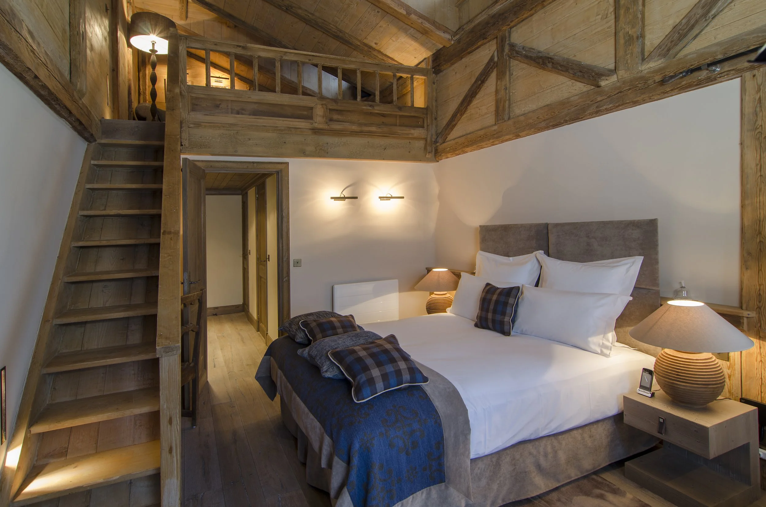 Schlafzimmer mit Holzdecke und Möbel, Doppelbett mit weißen Kissen und Bettdecke, Nachttische mit Lampen, obere Holzgalerie, Treppe, gemütliche Beleuchtung, gemütliches Interieur.