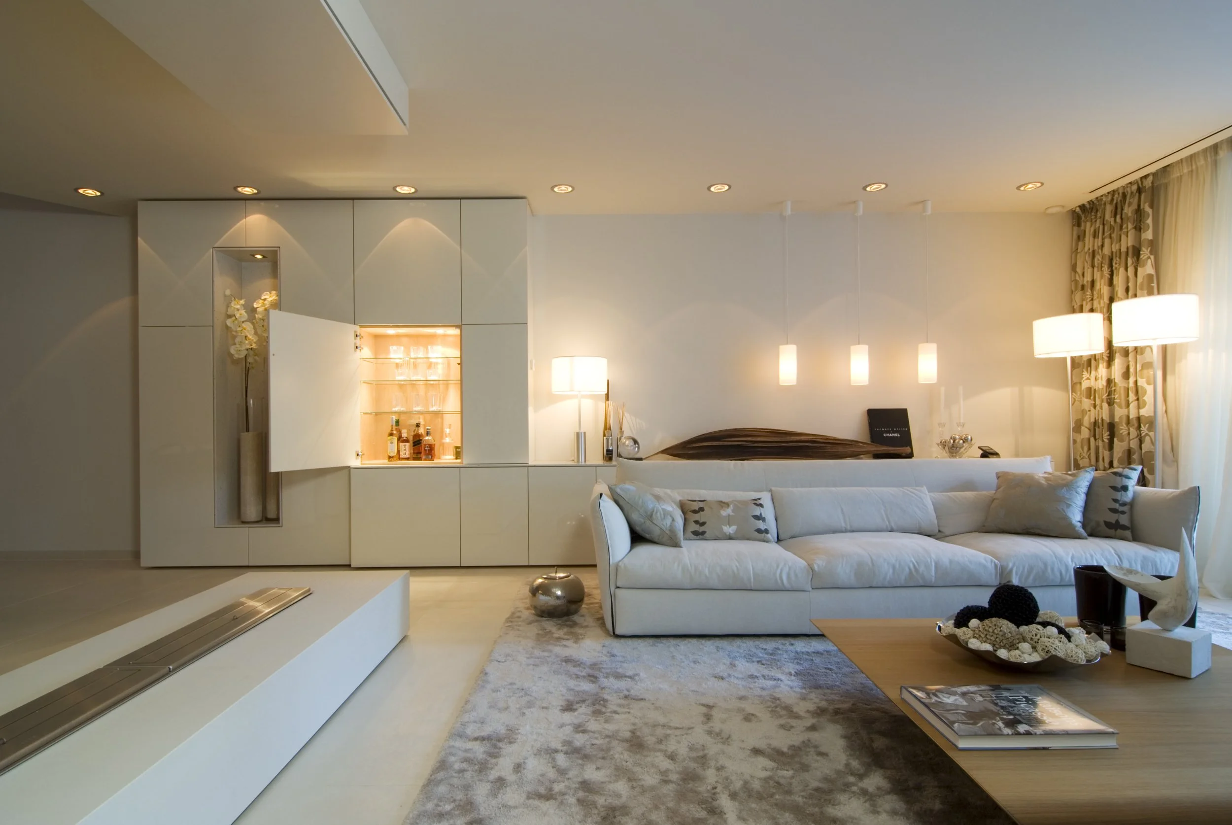 Moderne helle Wohnzimmer mit weißen Sofa, Couchtisch, dekorativen Kissen, stehendem Bodenleuchte und Tischlampen, Flachbildfernseher, Kamin und Gardinen mit Blumenmuster an großen Fenstern.