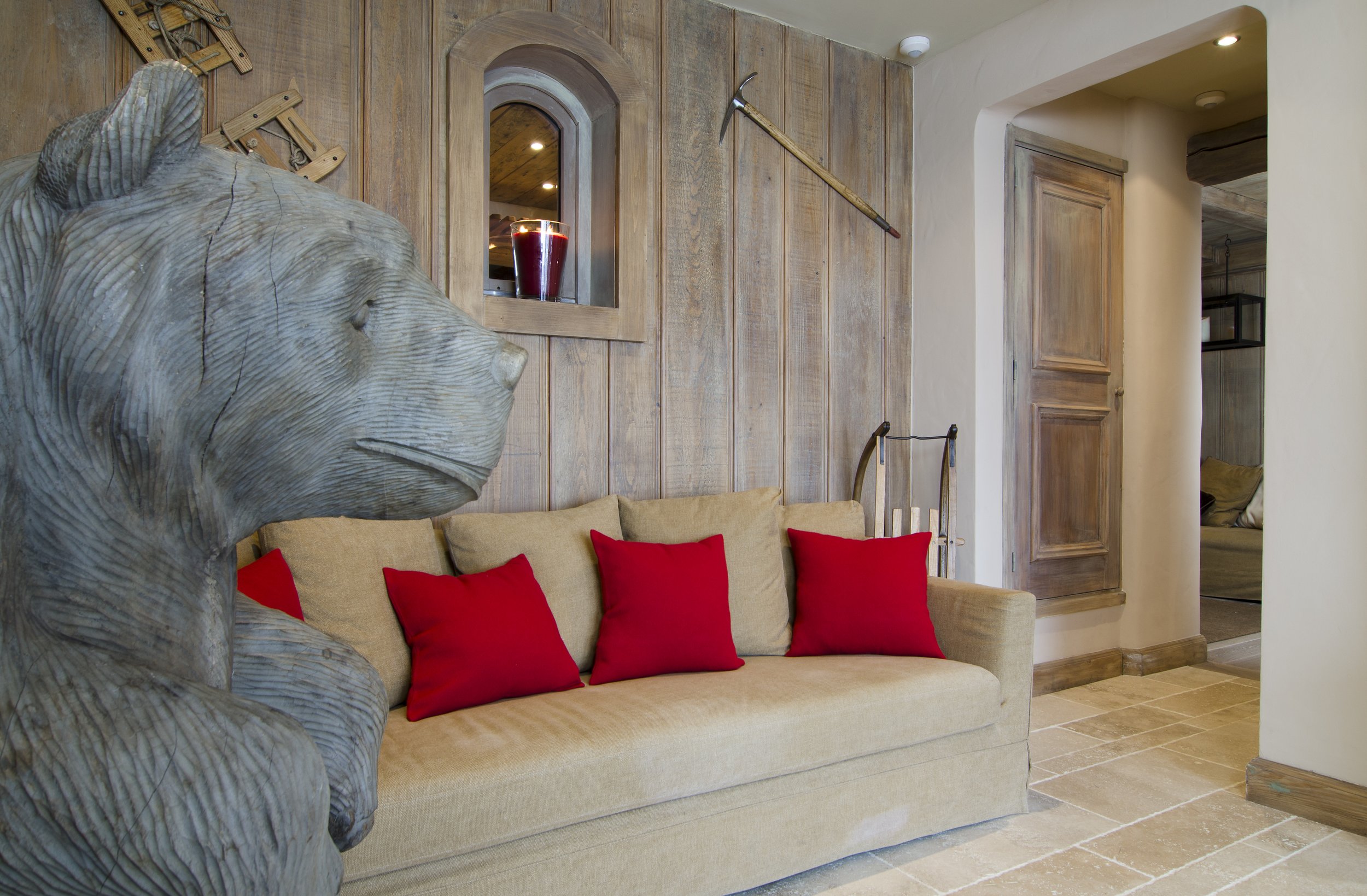 Ein Wohnzimmer mit einer beige Couch, roten Kissen und hölzernen Wandverkleidungen. Es gibt eine Skulptur eines Löwenkopfs im Vordergrund, sowie eine Fensteröffnung mit einer Kerze. An der Wand hängen landwirtschaftliche Werkzeuge, darunter eine Sens