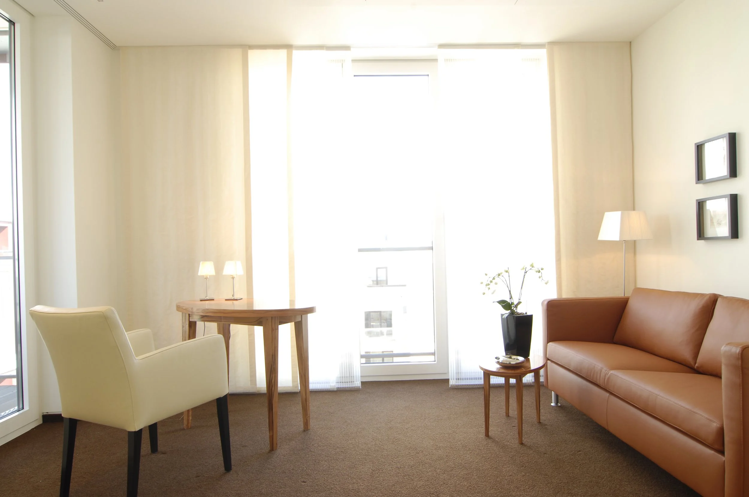 Helles Wohnzimmer mit großen Fenstern, brauner Teppich, braunes Sofa, weiße und cremefarbene Möbel, kleine Tischlampe, Stehlampe, Topfpflanze, Wandbilder.