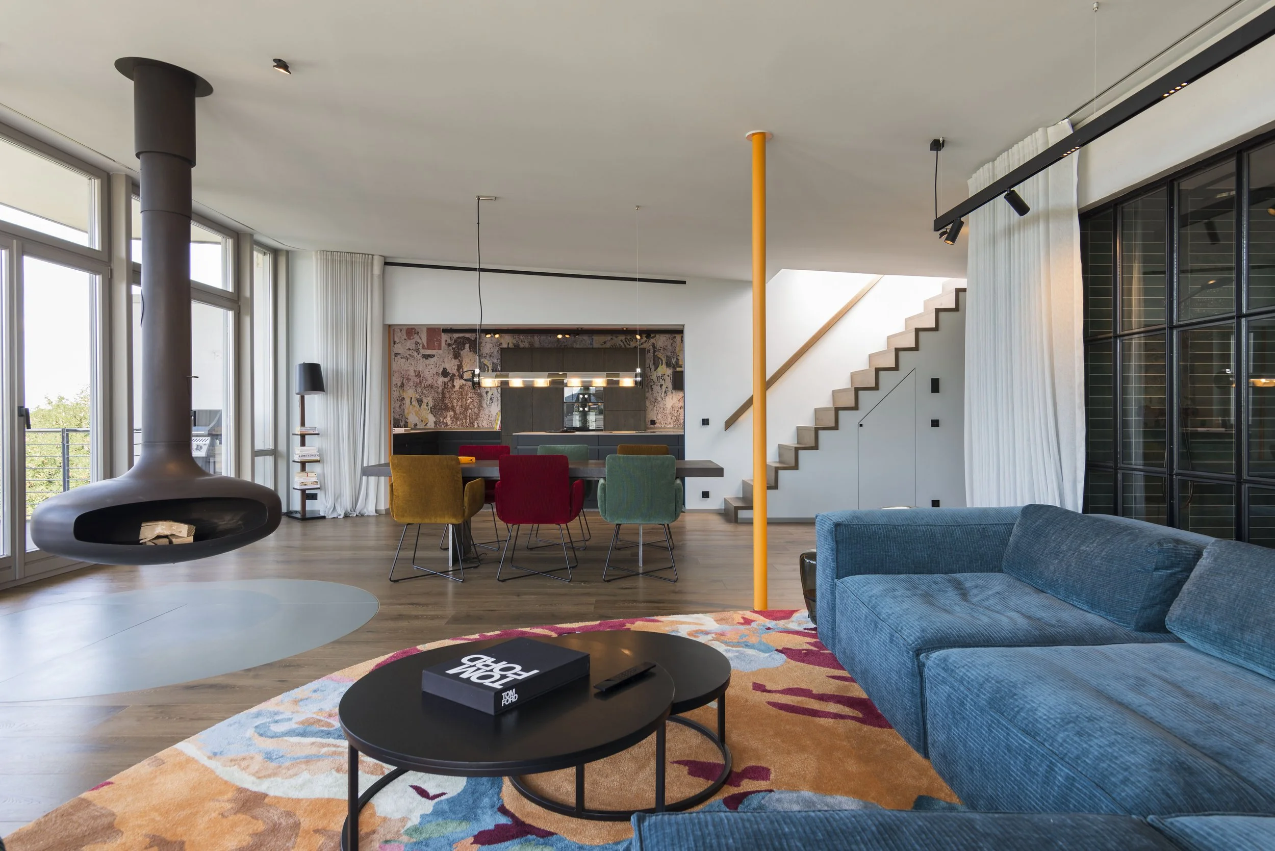 Modernes Wohnzimmer mit blauer Couch, farbigem Teppich, Kamin, offener Essbereich mit bunten Stühlen, Treppe und großen Fenstern, die viel Tageslicht lassen.
