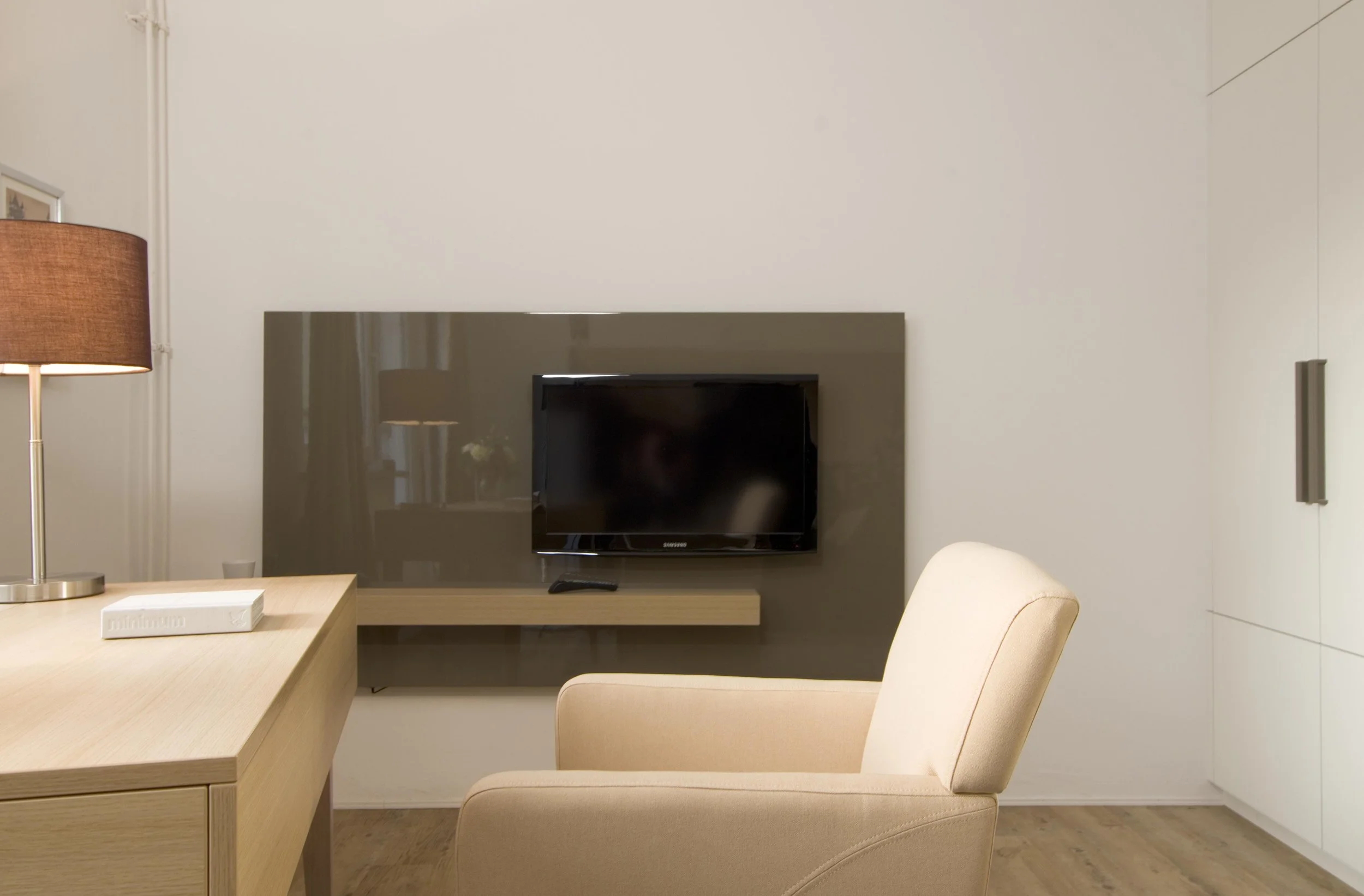 Moderner Wohnraum mit beige Sessel, dunklem Fernseher, Holzkommode, Tischlampe und weißer Wand.