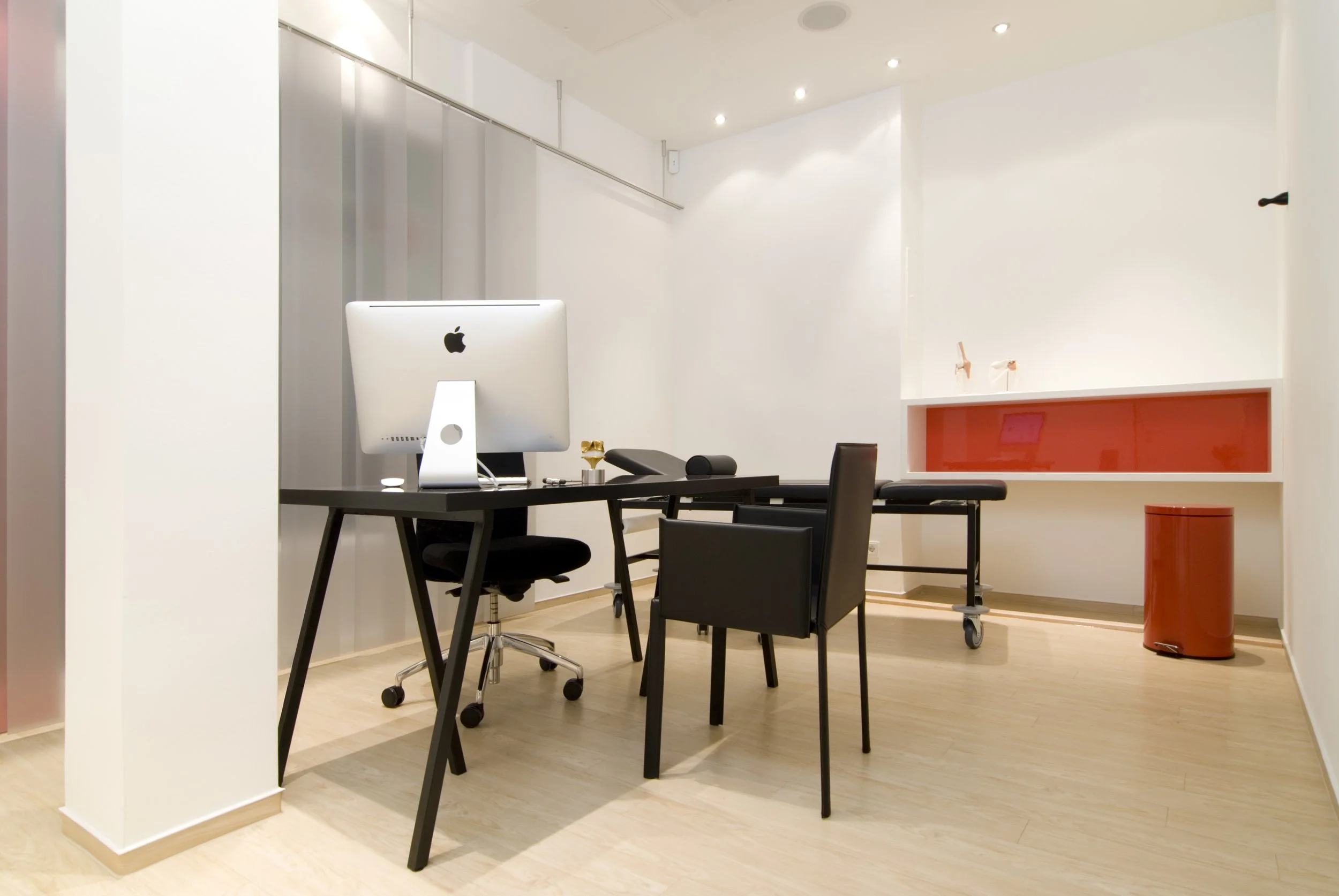 Ein modernes, minimalistisch gestaltetes Behandlungszimmer mit einem Schreibtisch, einem iMac-Computer, drei schwarzen Stühlen, einem Behandlungsstuhl, einem roten Abfalleimer und einem roten Ablagefach an der Wand.