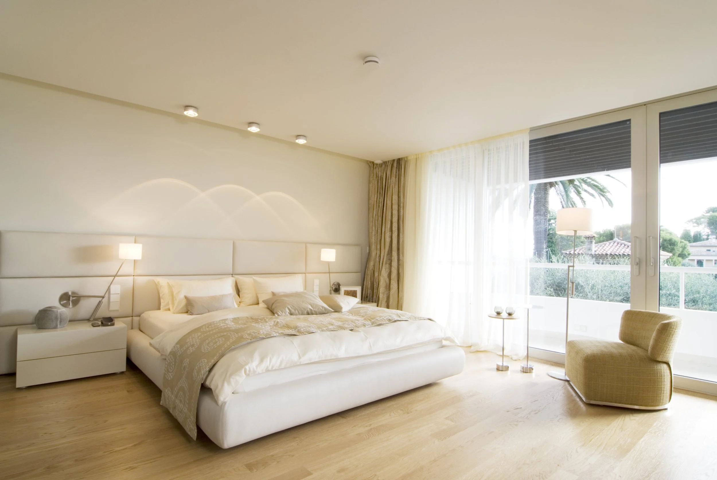 Ein helles, modernes Schlafzimmer mit einem großen Bett, seitlichen Nachttischen, Stehlampen, einem Sessel, einem kleinen Beistelltisch und großen Fenstern mit Blick nach draußen auf Palmen und einen Garten.