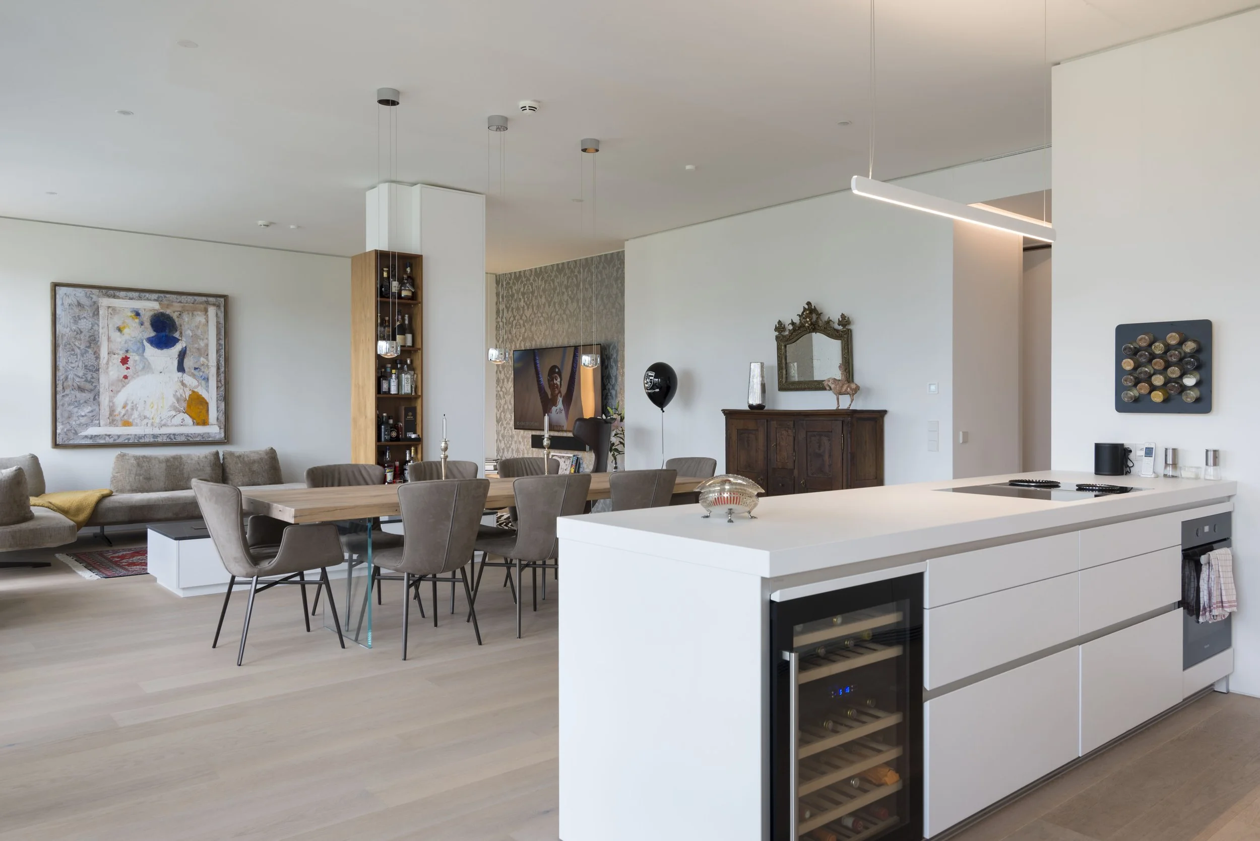 Offene Wohnküche mit weißem Kühl- und Kochblock im Vordergrund, Wohnzimmer mit Esstisch, Sesseln und Wandkunst im Hintergrund.