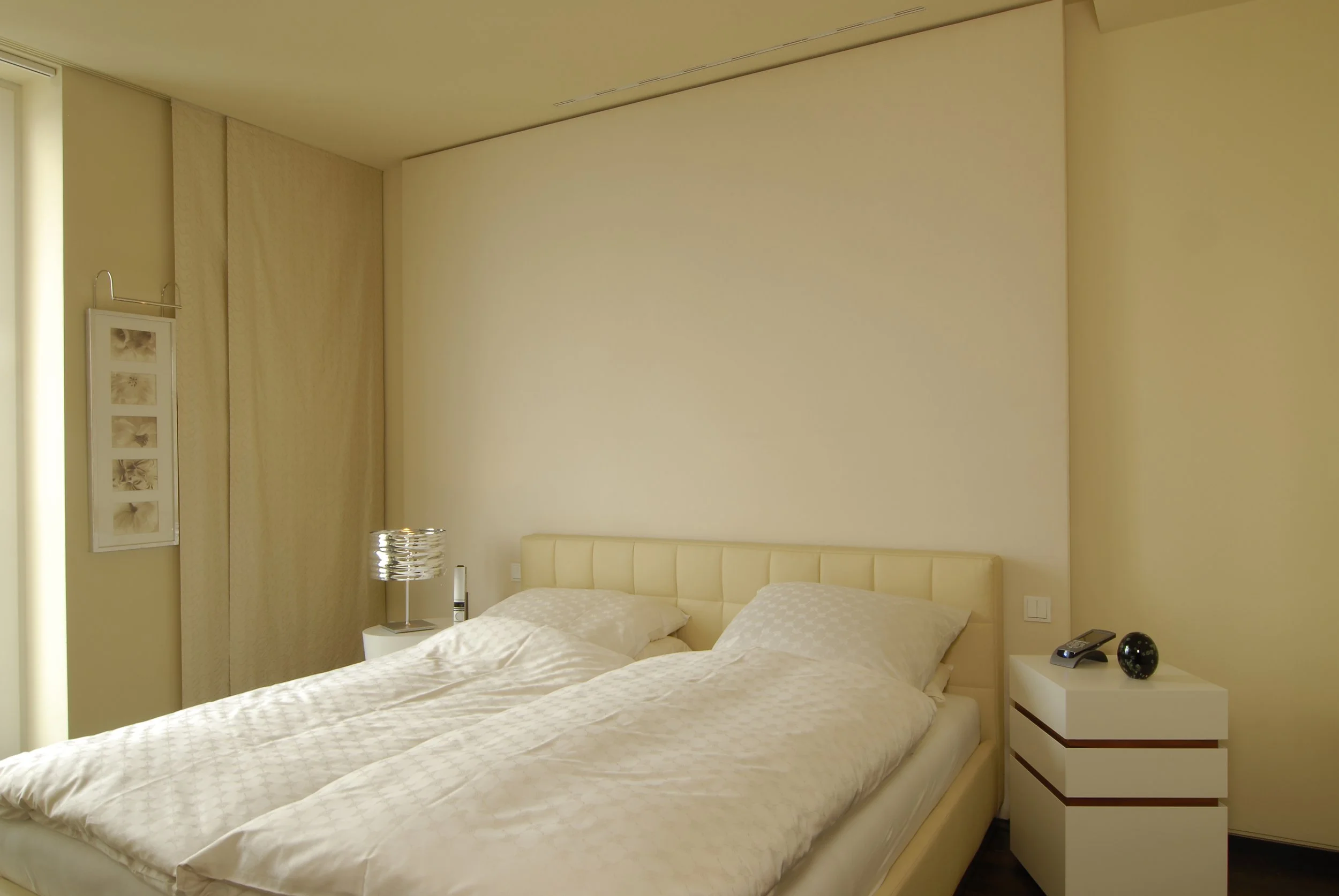 Ein Schlafzimmer mit einem großen Doppelbett, das mit weißer Bettwäsche bedeckt ist, zwei Nachttische, one with einen modernen Lampen, das andere mit einem Telefon und einer dekorativen schwarzen Schale, Wanddekoration mit Fotos, beige Vorhänge, schl