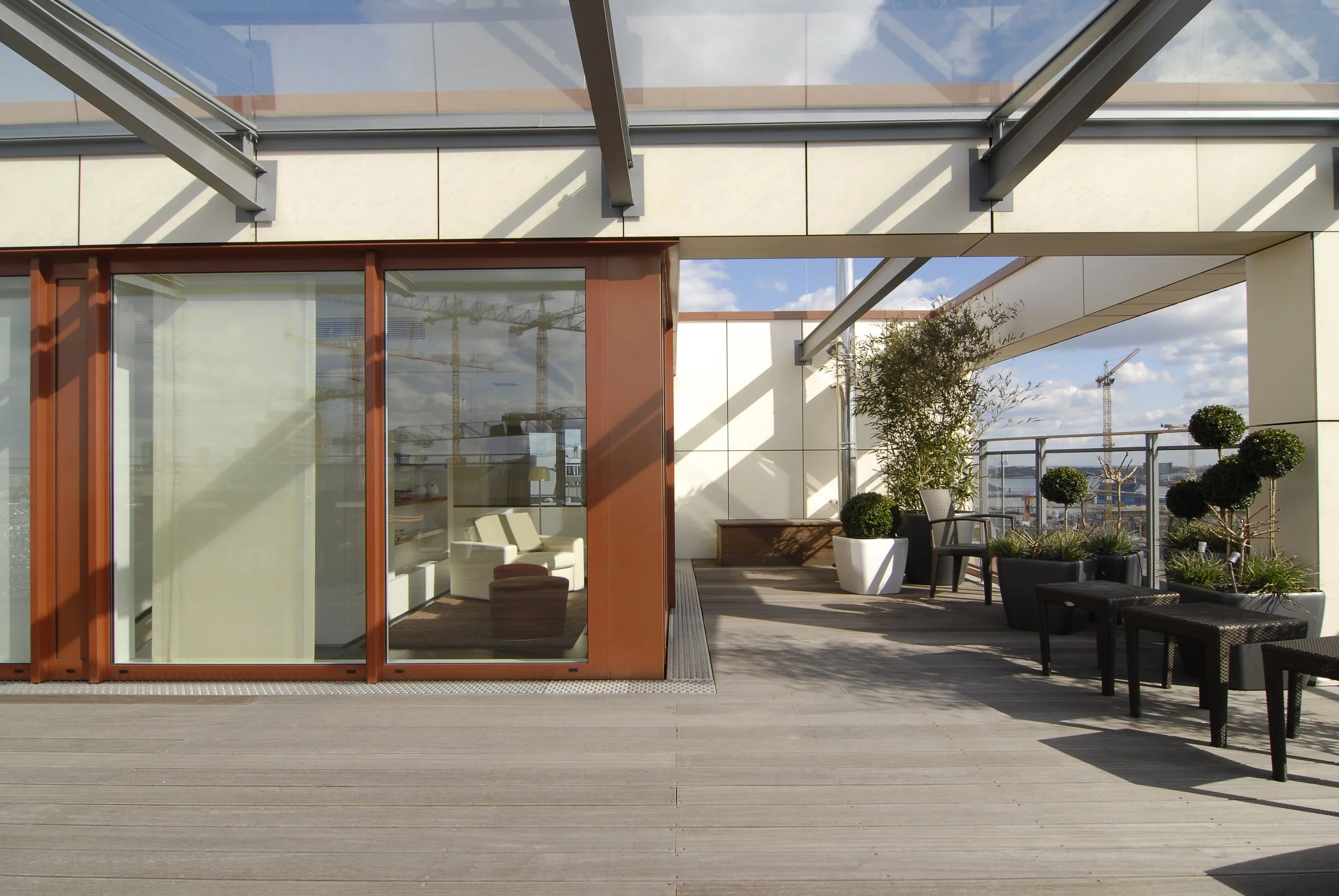 Blick auf eine moderne Dachterrasse mit Sitzgelegenheiten und pflanzen, umgeben von Glas, Holz und Metallstrukturen. Im Hintergrund sind Baukräne und eine Stadt zu sehen.