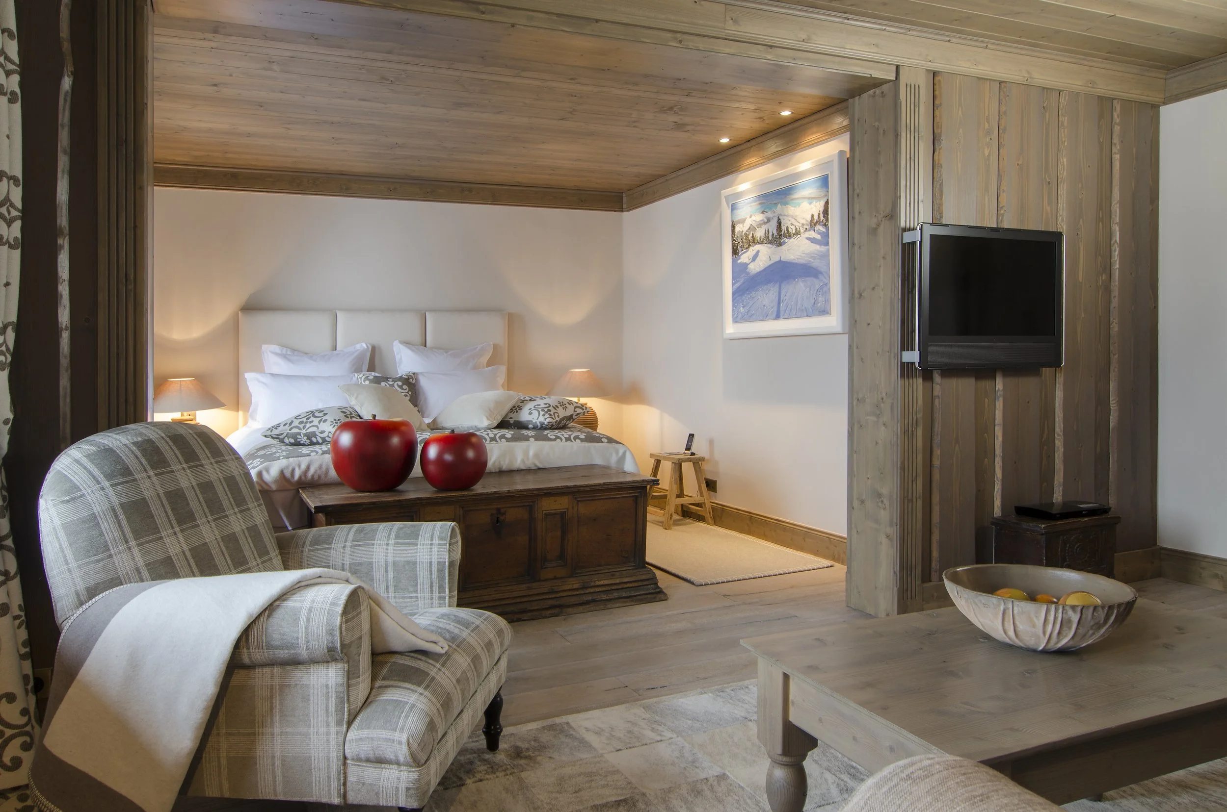 Rustikale Hotelzimmer mit Holzwand und -decke, Komfortbett mit weißen und gemusterten Kissen, Nachtische mit Lampen, Fenster mit Schneelandschaft, Sessel mit Decke, Fernseher, Couchtisch mit Obstschale.