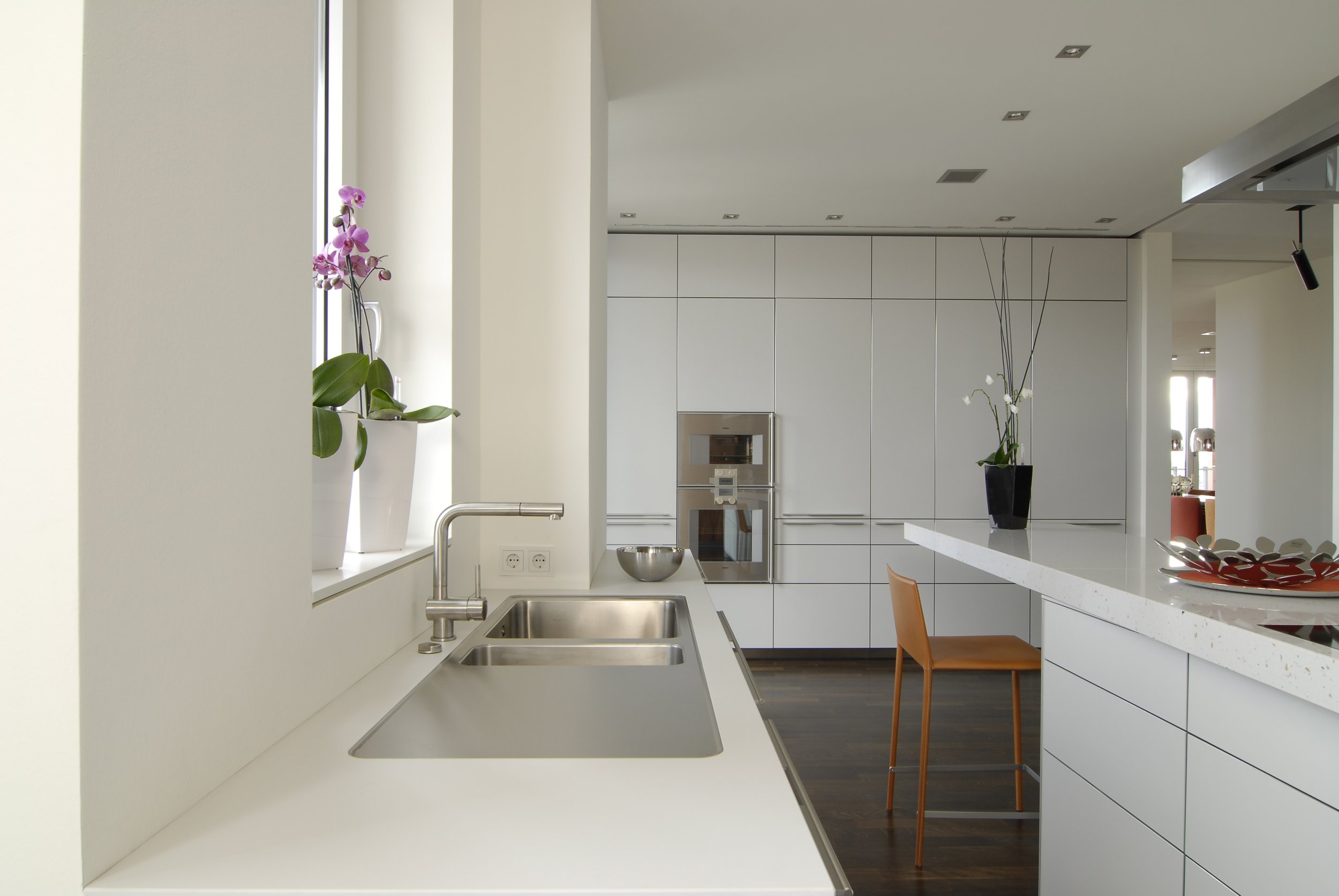 Moderne weiße Küche mit Doppelspüle, pflanzen auf Fensterbank und hoher weißer Schrankwand im Hintergrund, brauner Holzstuhl am weißen Küchenblock mit Marmoroptik.