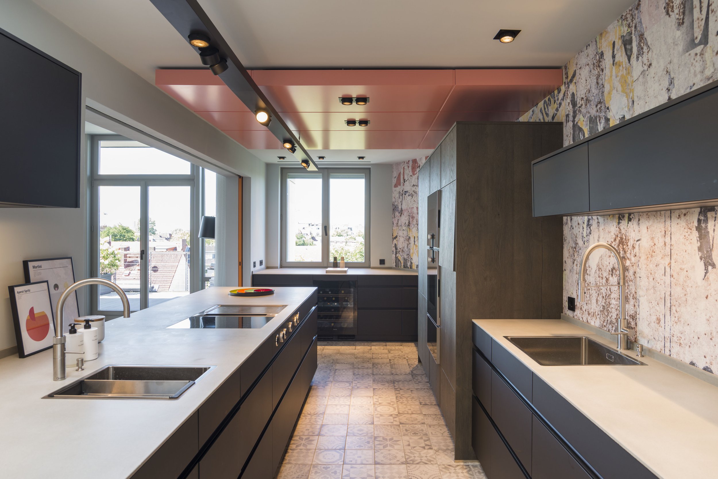Moderne Küche mit schwarzen Schränken, weißer Arbeitsplatte, two Spülbecken, großen Fenstern, rustikalem Wanddesign, beleuchtet mit Deckenleuchten und einer Deckenlampe, Blick nach draußen auf einen Garten