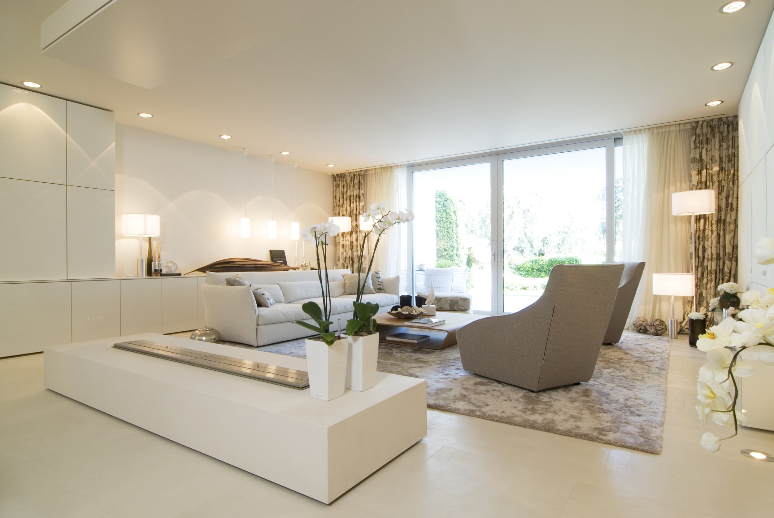 Moderner, heller Wohnraum mit weißen Möbeln, großen Fenstern und Blick auf einen Garten mit Bäumen und Sträuchern.