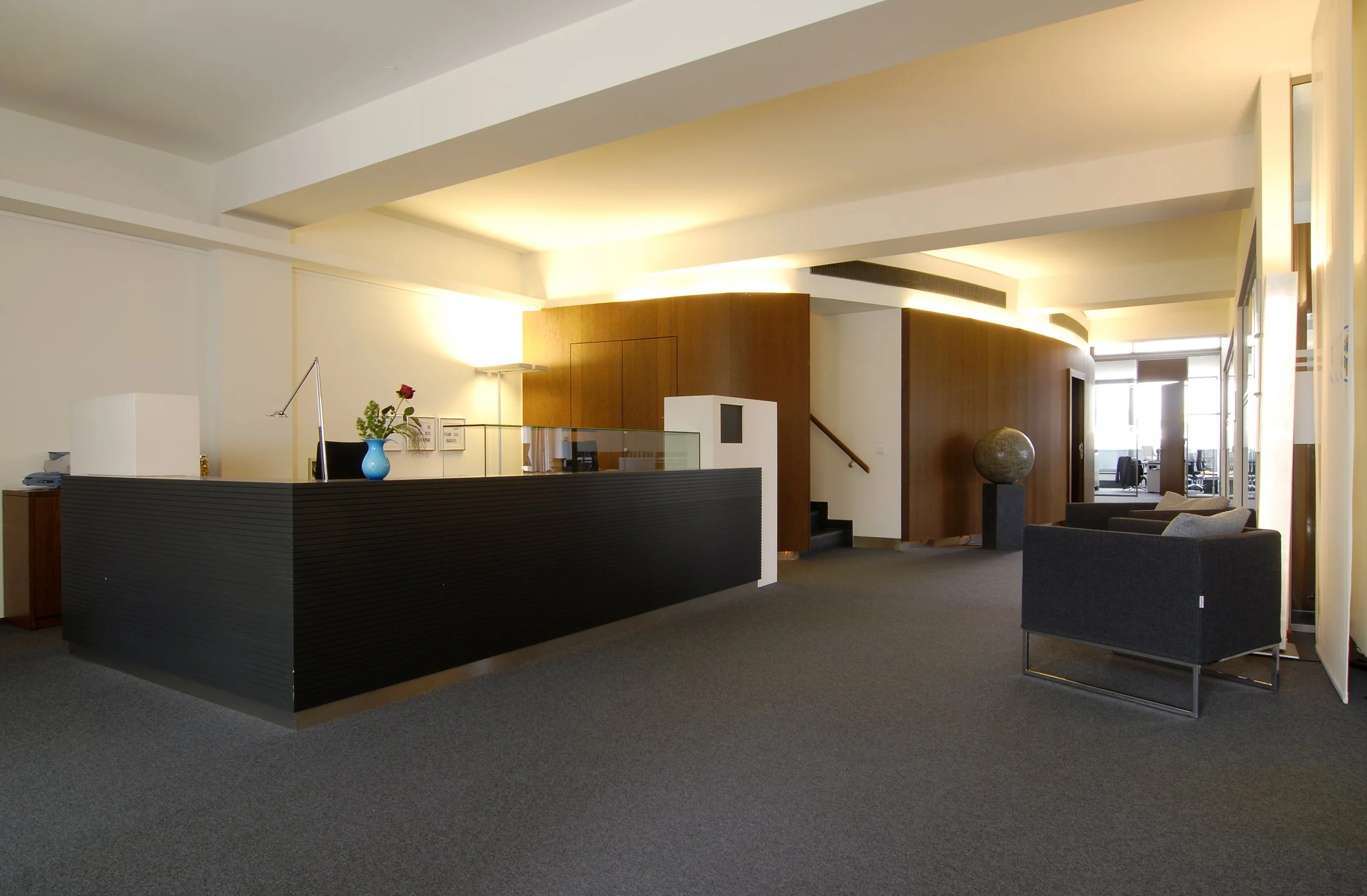 Moderner Hotelempfangsbereich mit schwarzer Rezeption, Stehpult, grauem Teppich, Skulptur, Sitzbereich und Holzverkleidung an der Wand.
