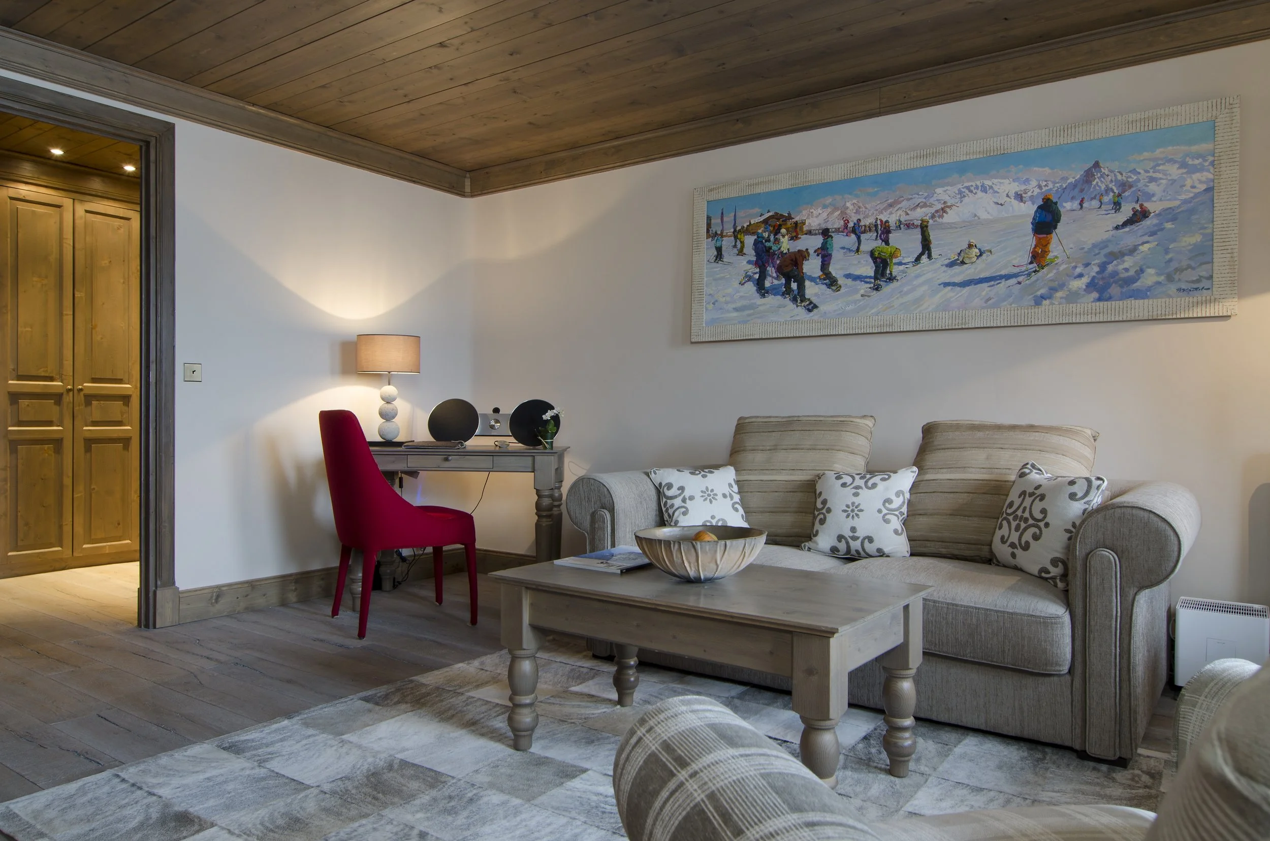 Gemütliches Wohnzimmer mit hellem Sofa, Kissen, Couchtisch, Wandbild von Skifahrern im Schnee, Schreibtisch mit Lampe, roter Stuhl und Holztüren im Hintergrund.