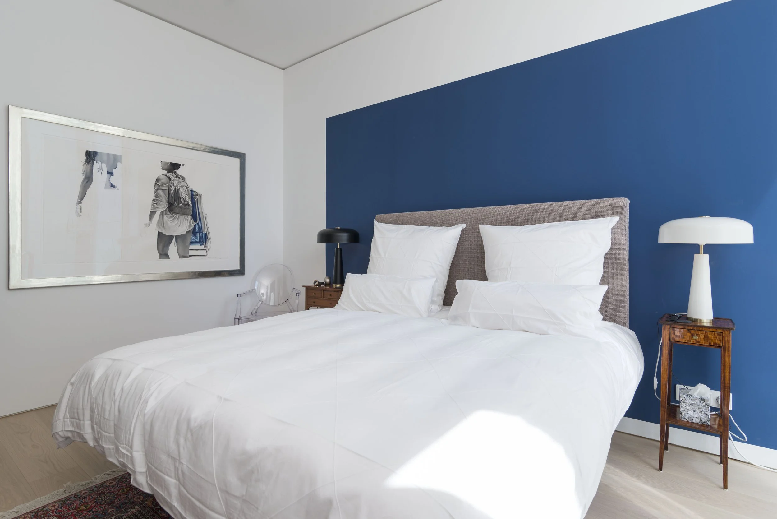 Schlafzimmer mit weißem Bett, blauer Wand im Hintergrund, zwei Nachttische mit Lampen, großes Wandbild mit schwarzen und weißen Fotos.