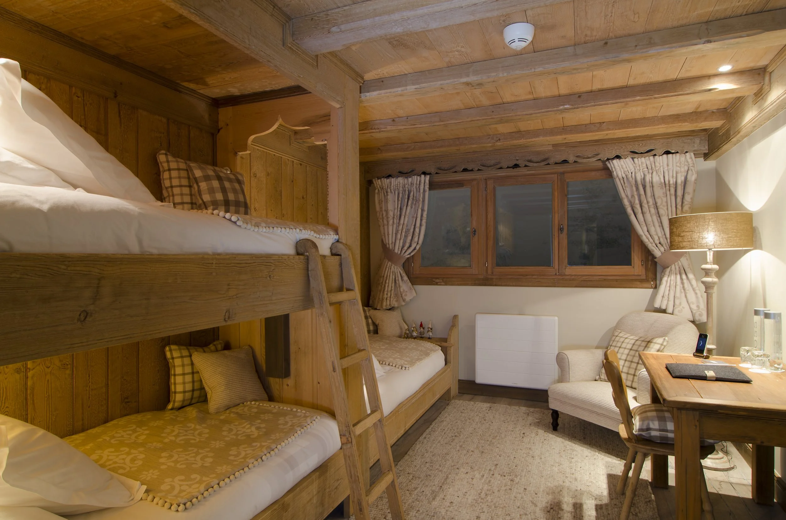 Gemütliches Schlafzimmer mit Holz-Doppel- und Einzelbetten, großen Fenstern mit Vorhängen, beigem Sessel, Holzschreibtisch mit Stuhl,Stehlampe, Teppichboden, Holzdecke und -Wände.
